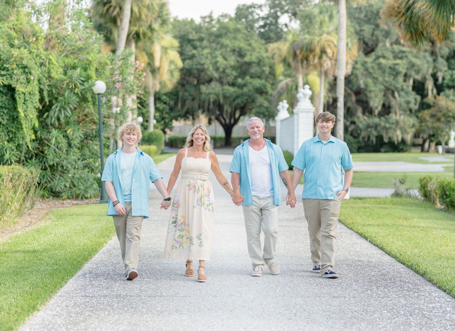 A family strolls together on a palm-lined path wearing coordinated light blue and white outfits in a tropical garden setting.