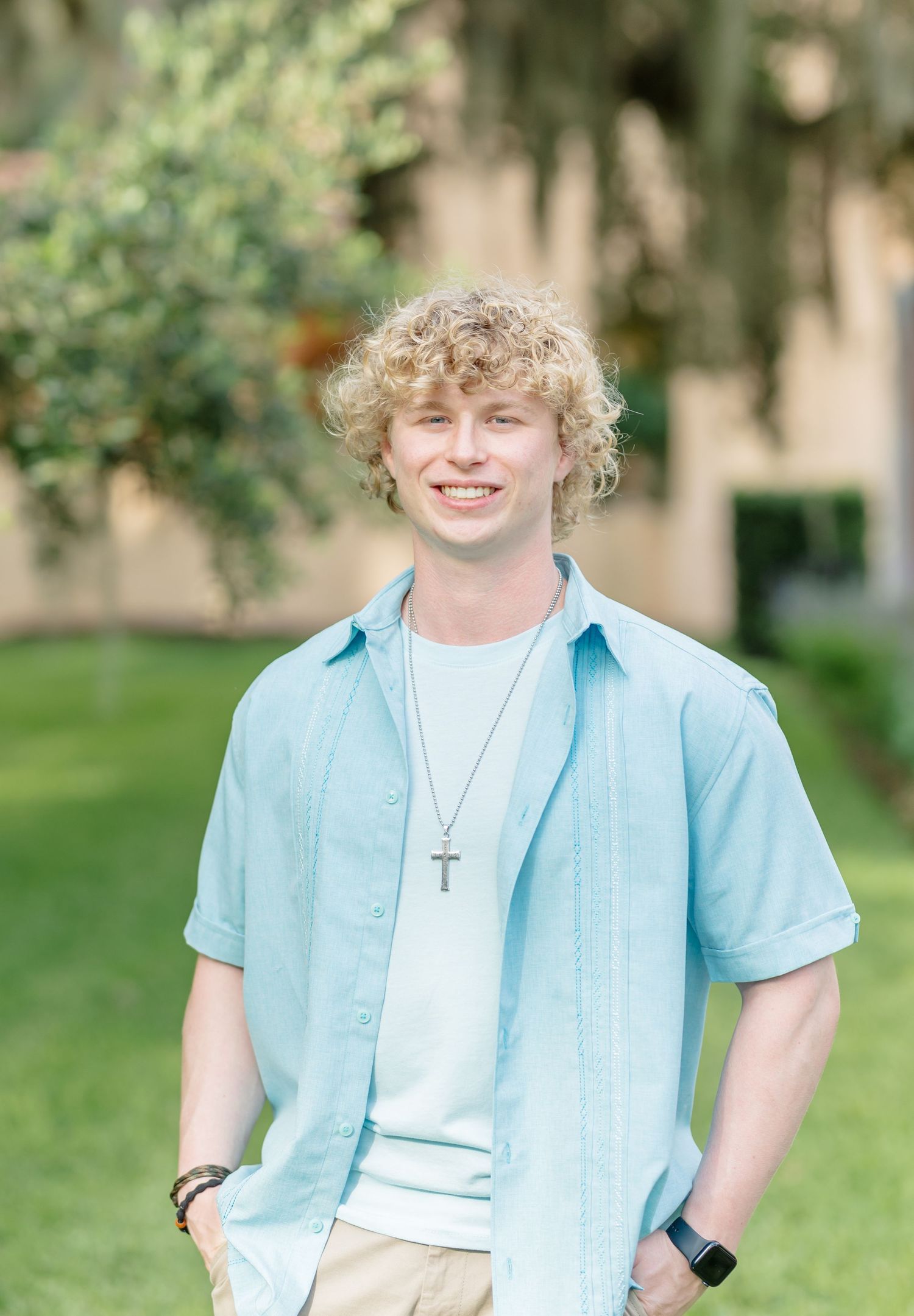 A young person in a light blue button-down shirt over a white t-shirt poses outdoors with a cross necklace.