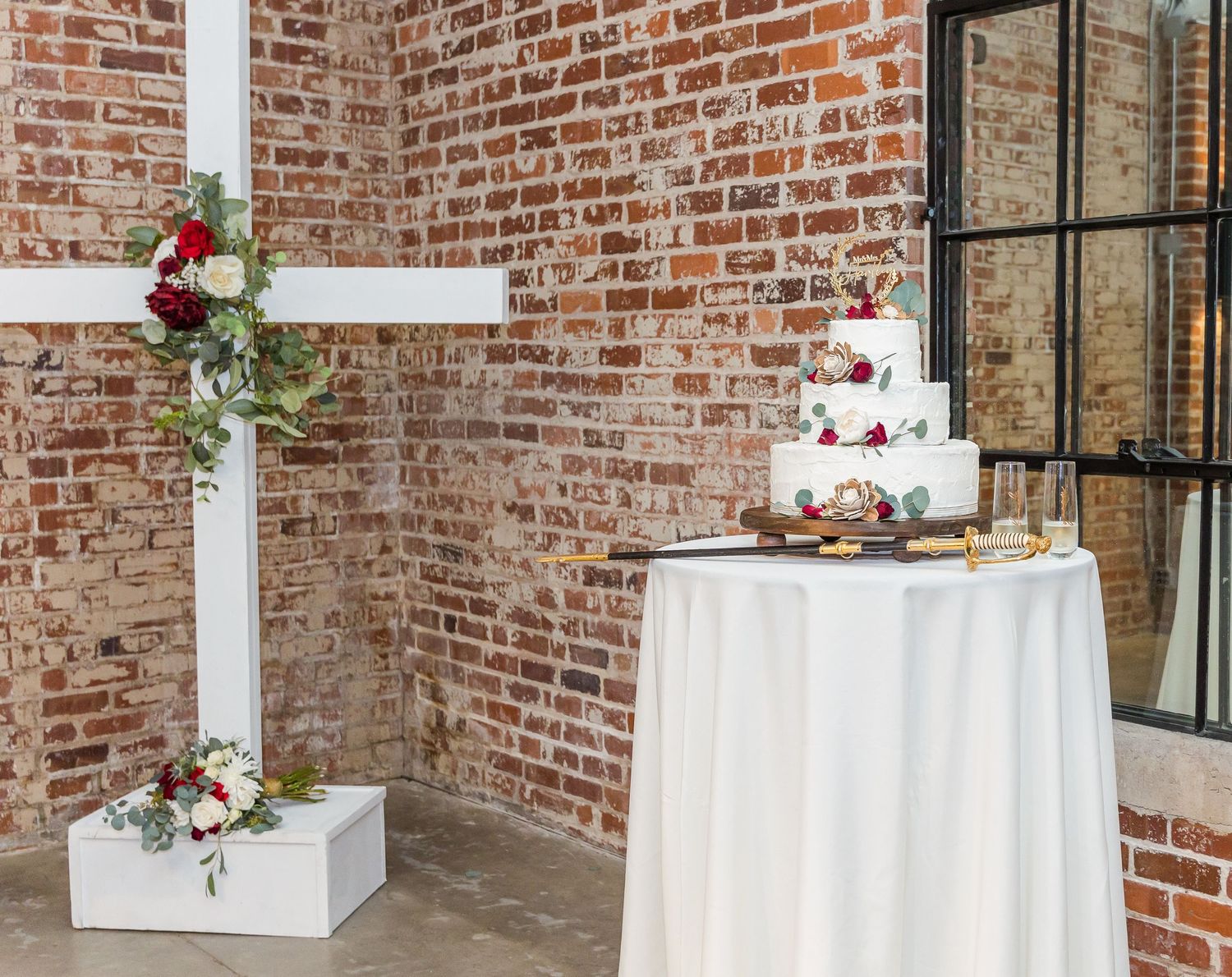 A white wedding cake sits on a table beside a rustic cross decorated with red and pink flowers against a brick wall.