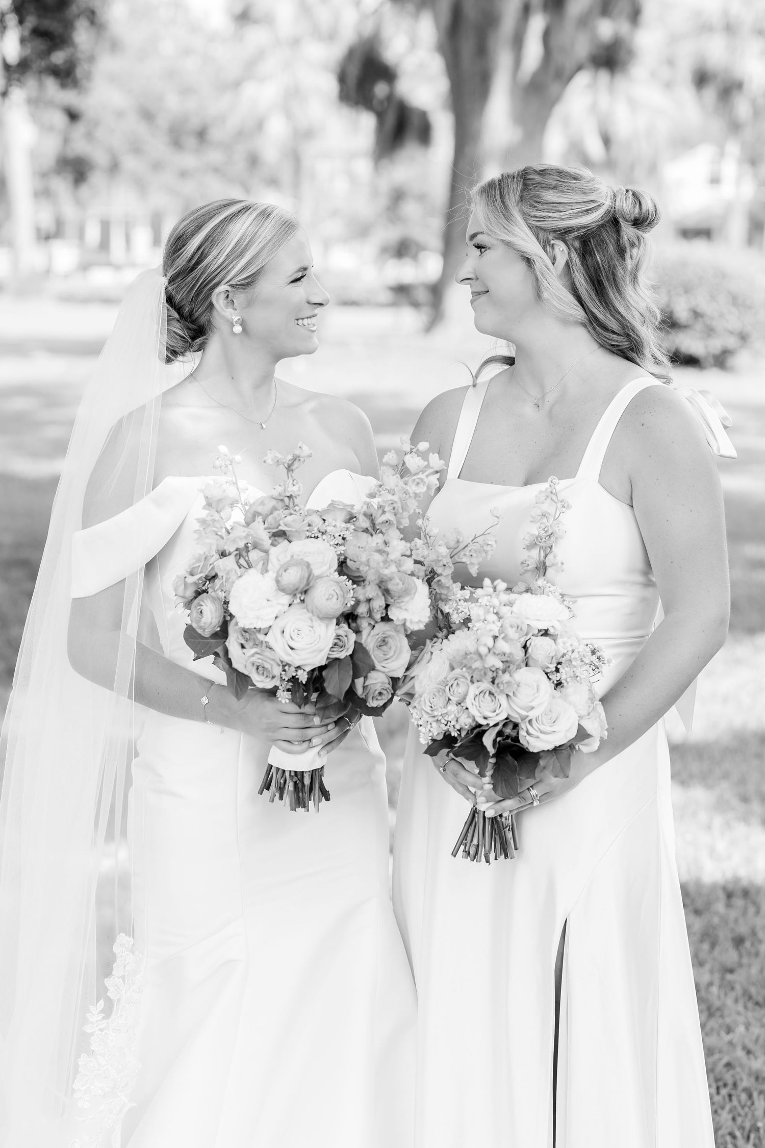 A black and white portrait of two attendants holding bouquets and sharing a candid moment.