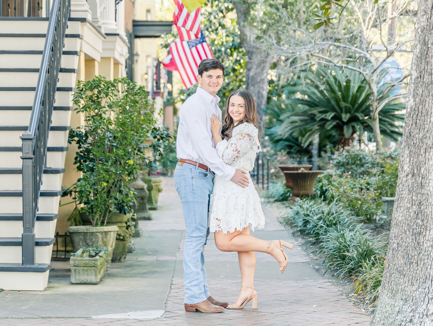 A couple embraces on a Charleston sidewalk with an American flag and lush greenery in the background.