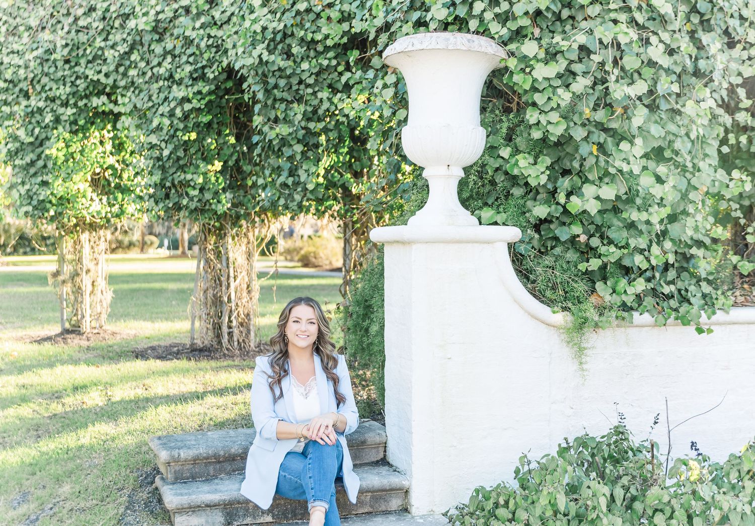A person in a white dress sits on a decorative stone bench in a lush garden setting.