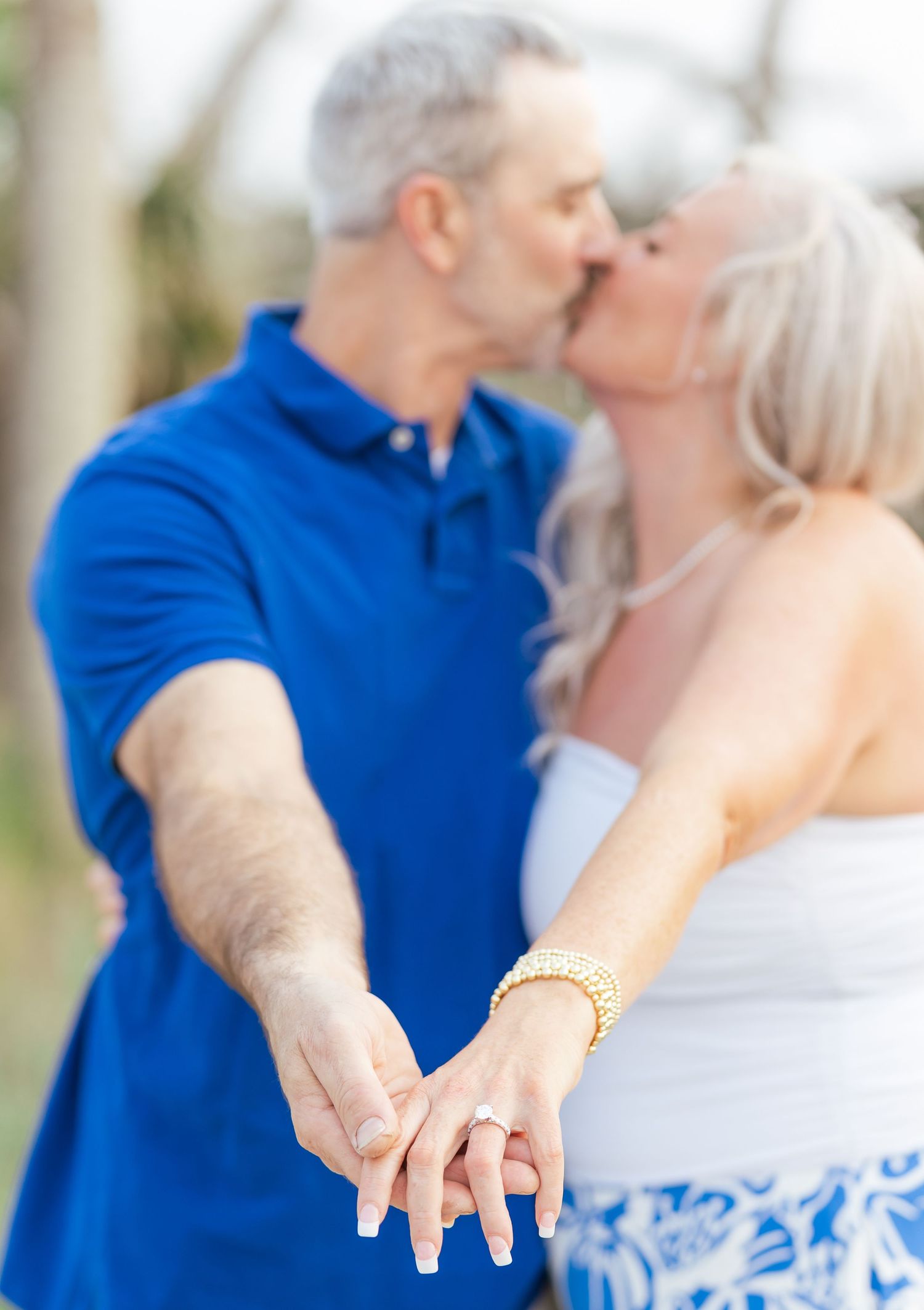 A romantic couple shares a kiss while showing off a wedding ring in a beach setting.