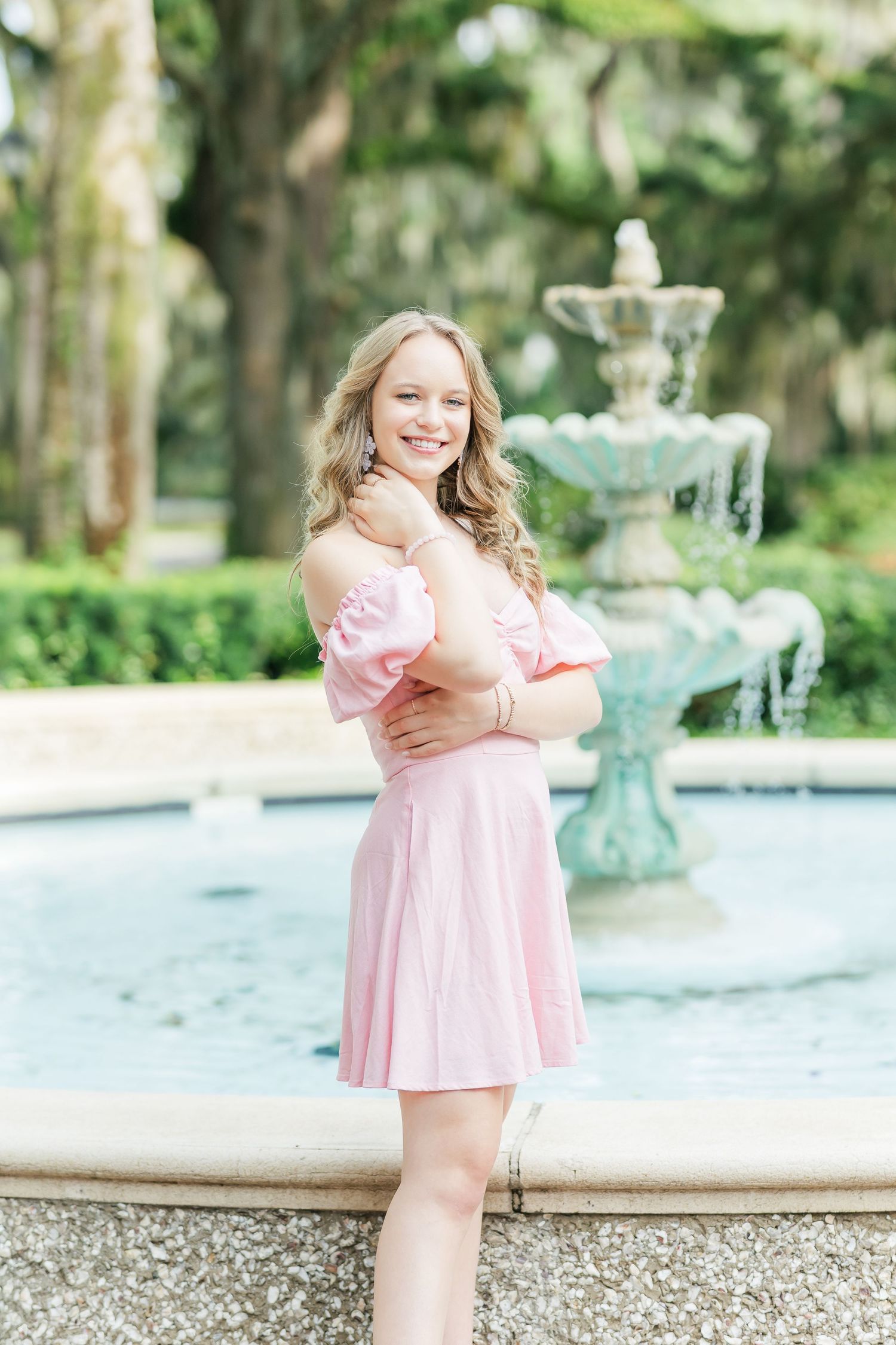 A pink dress-clad figure poses cheerfully by an ornate tiered fountain in a lush garden setting.