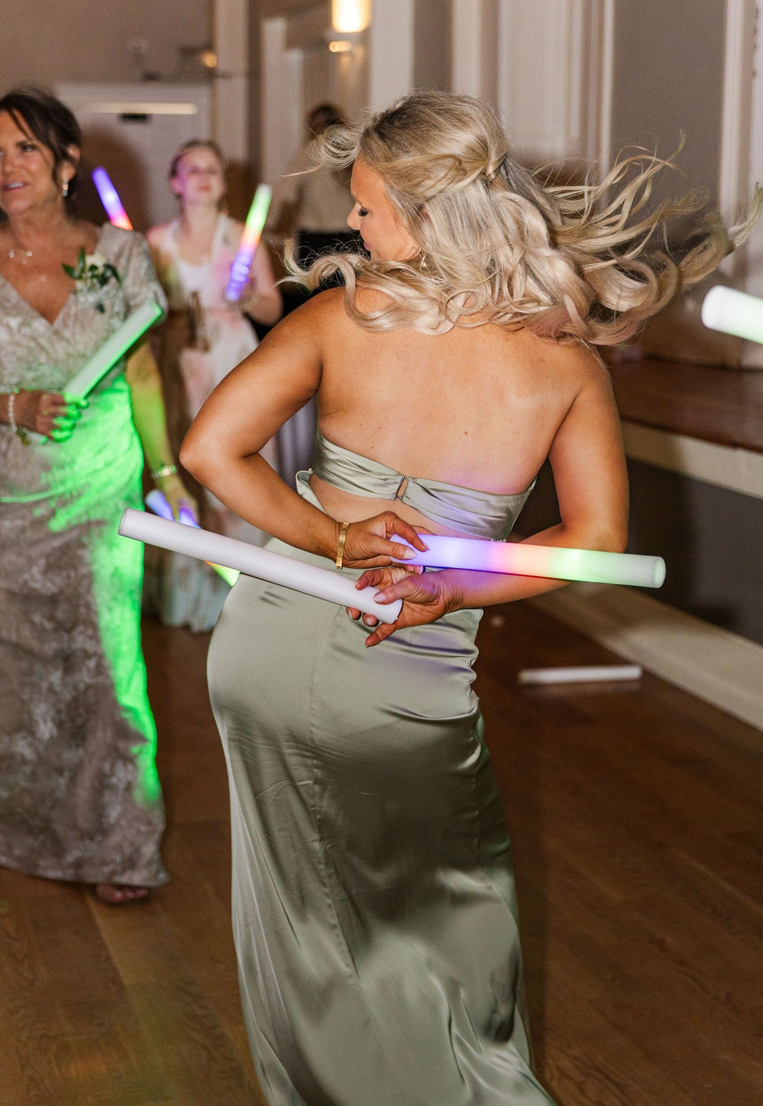 Bridesmaid in sage green gown twirls with glow sticks on wedding dance floor.