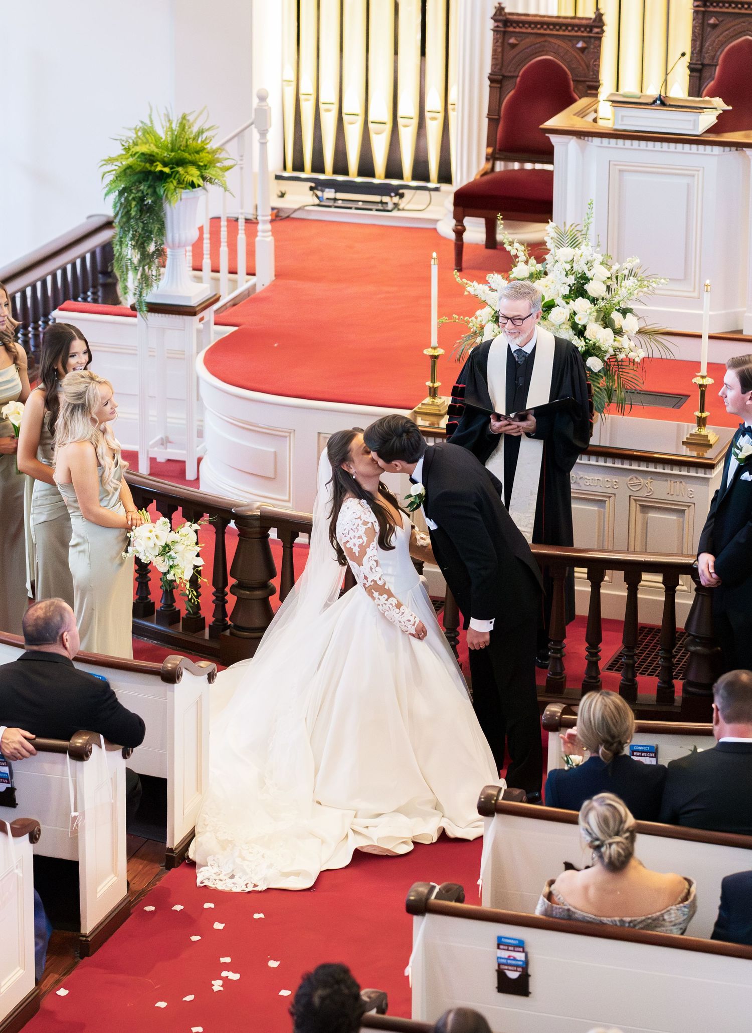 Bride and groom share first kiss at church wedding altar with officiant and bridesmaids nearby.