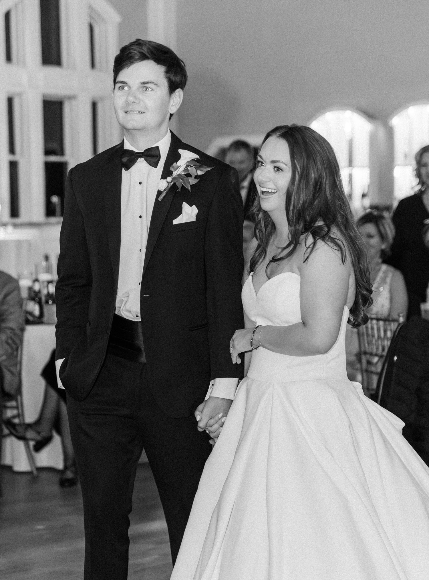 Black and white photo of a bride and groom holding hands and smiling as they enter their wedding reception.