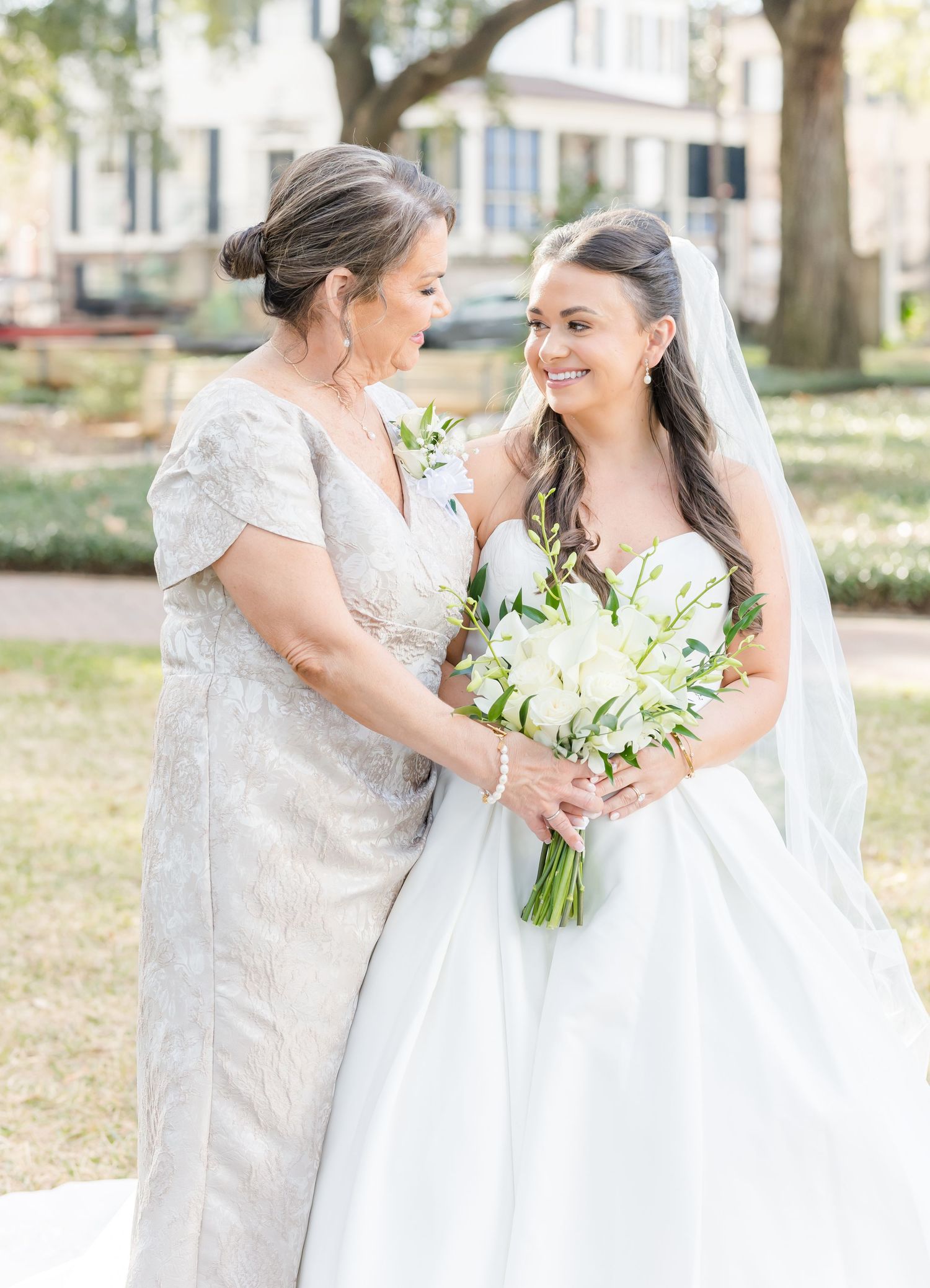 Smiling bride in white gown with veil holds white bouquet beside older woman in silver dress.