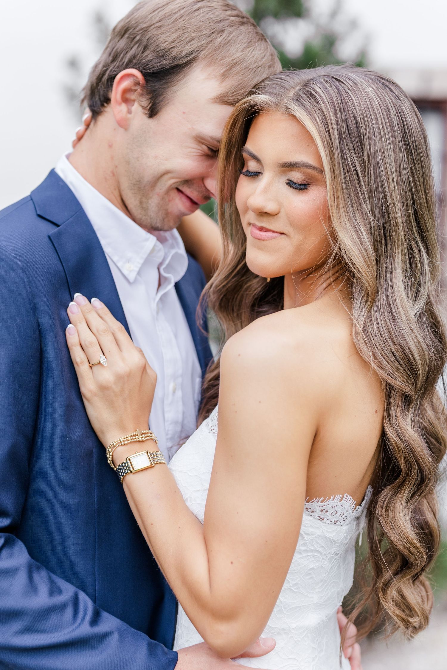 Couple in romantic embrace, woman in white lace dress with gold bracelet, man in navy blazer.
