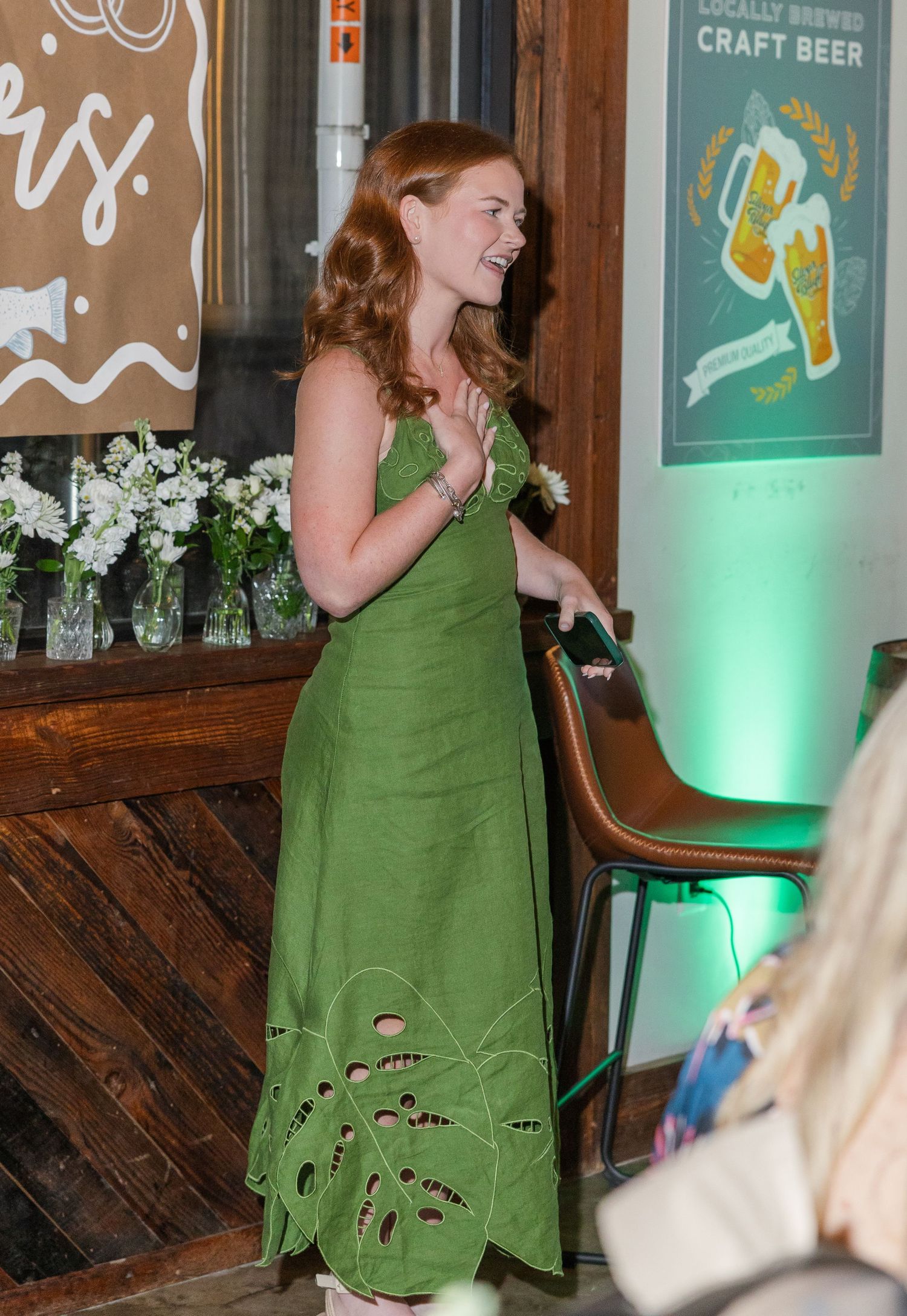 Woman in green floral midi dress smiling at craft beer event with white flower arrangements.
