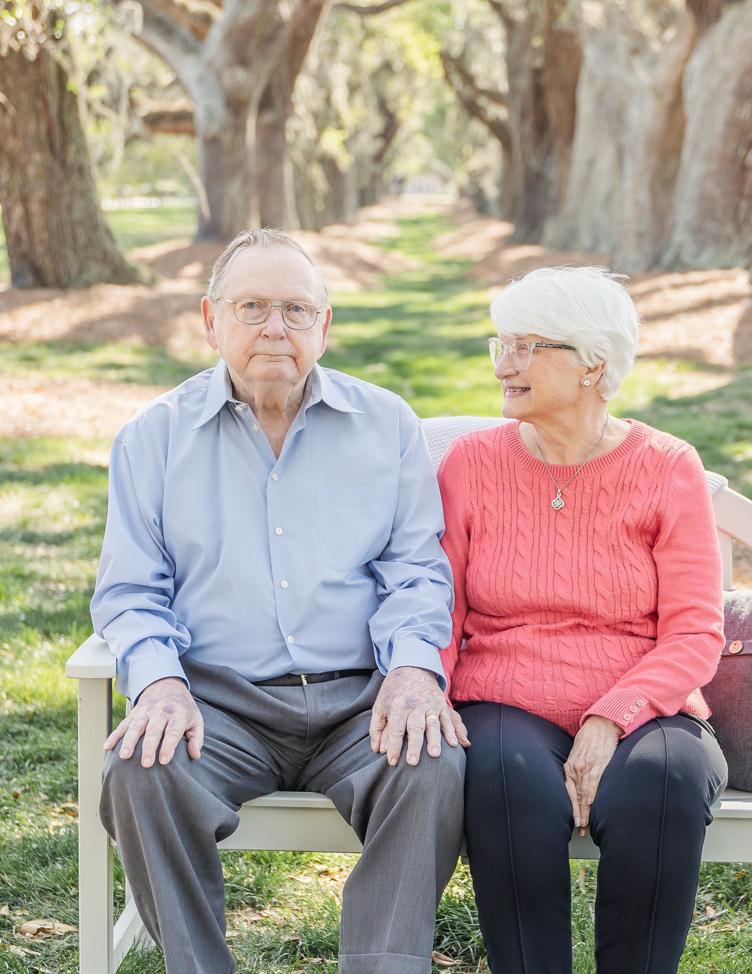 Senior couple sits close together on a wooden bench in a park with tree-lined path.