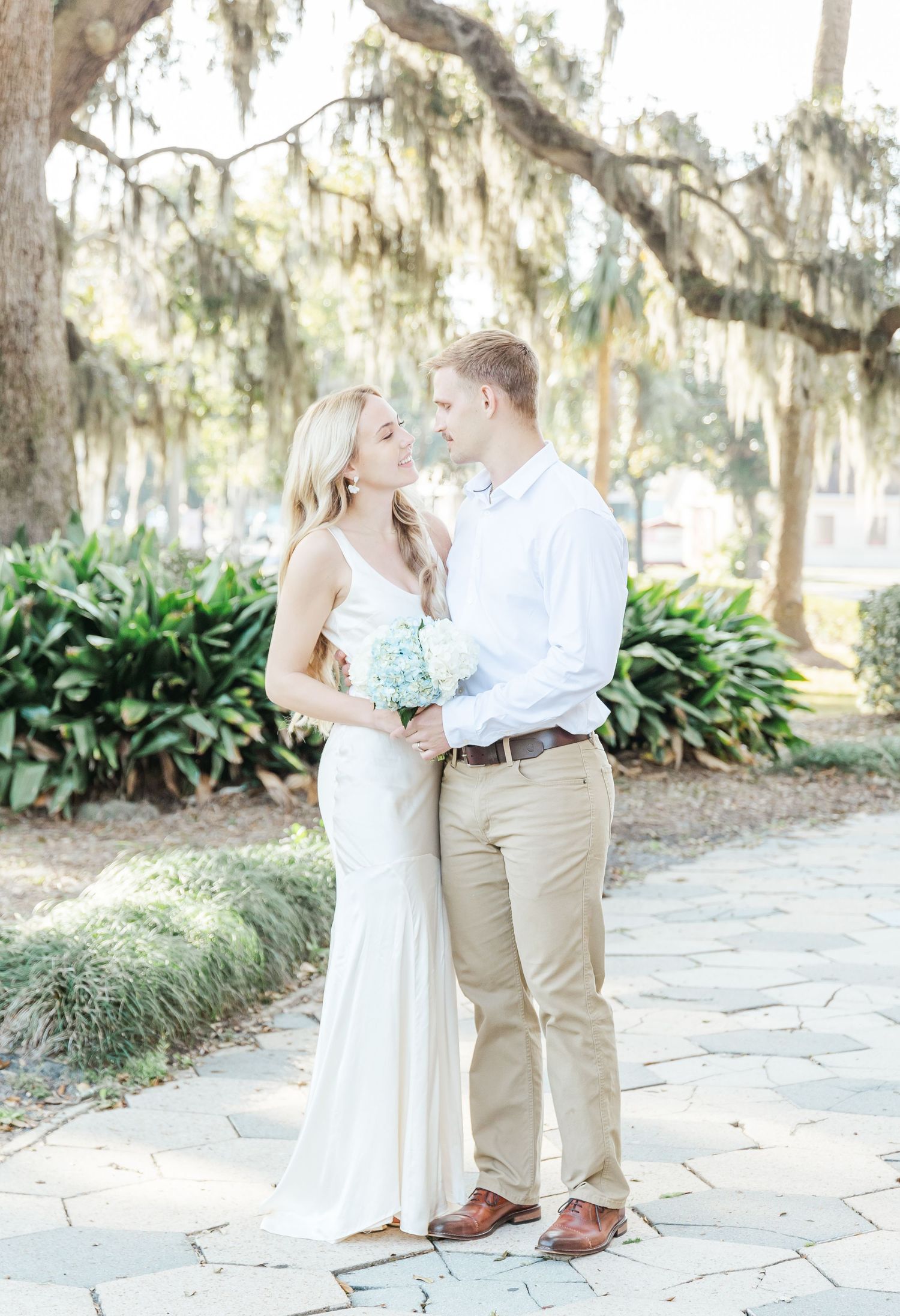 A romantic wedding couple poses under Spanish moss draped trees on a stone pathway beside tropical plants.