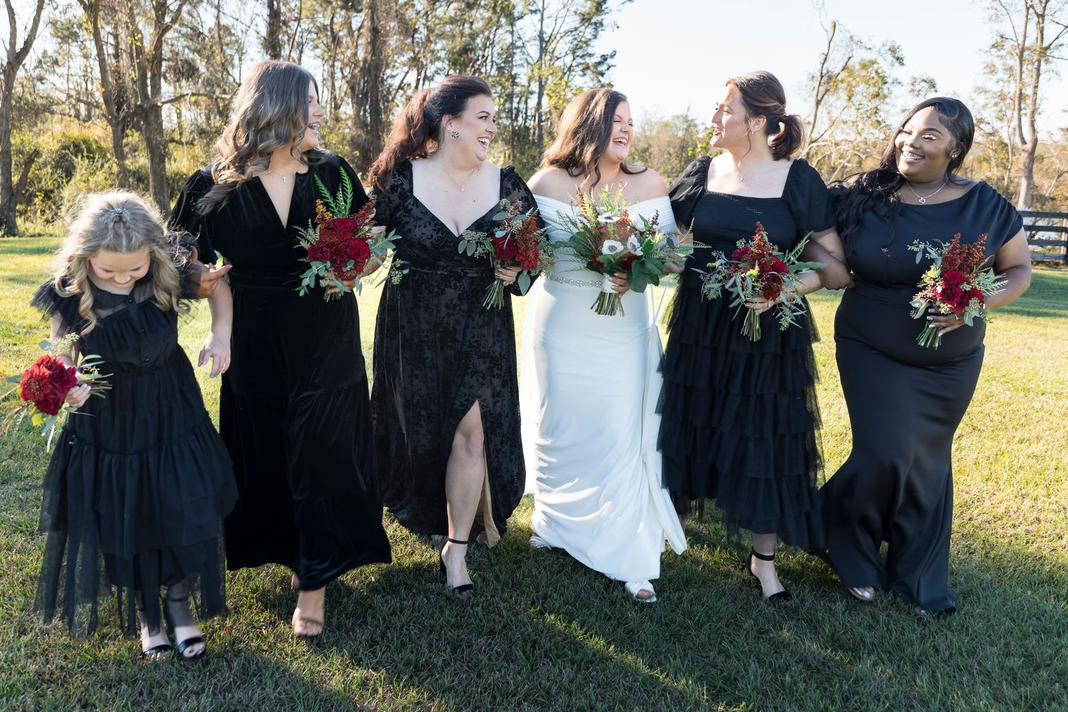 Wedding party in black dresses poses outdoors with bright red bouquets on a sunny autumn day.