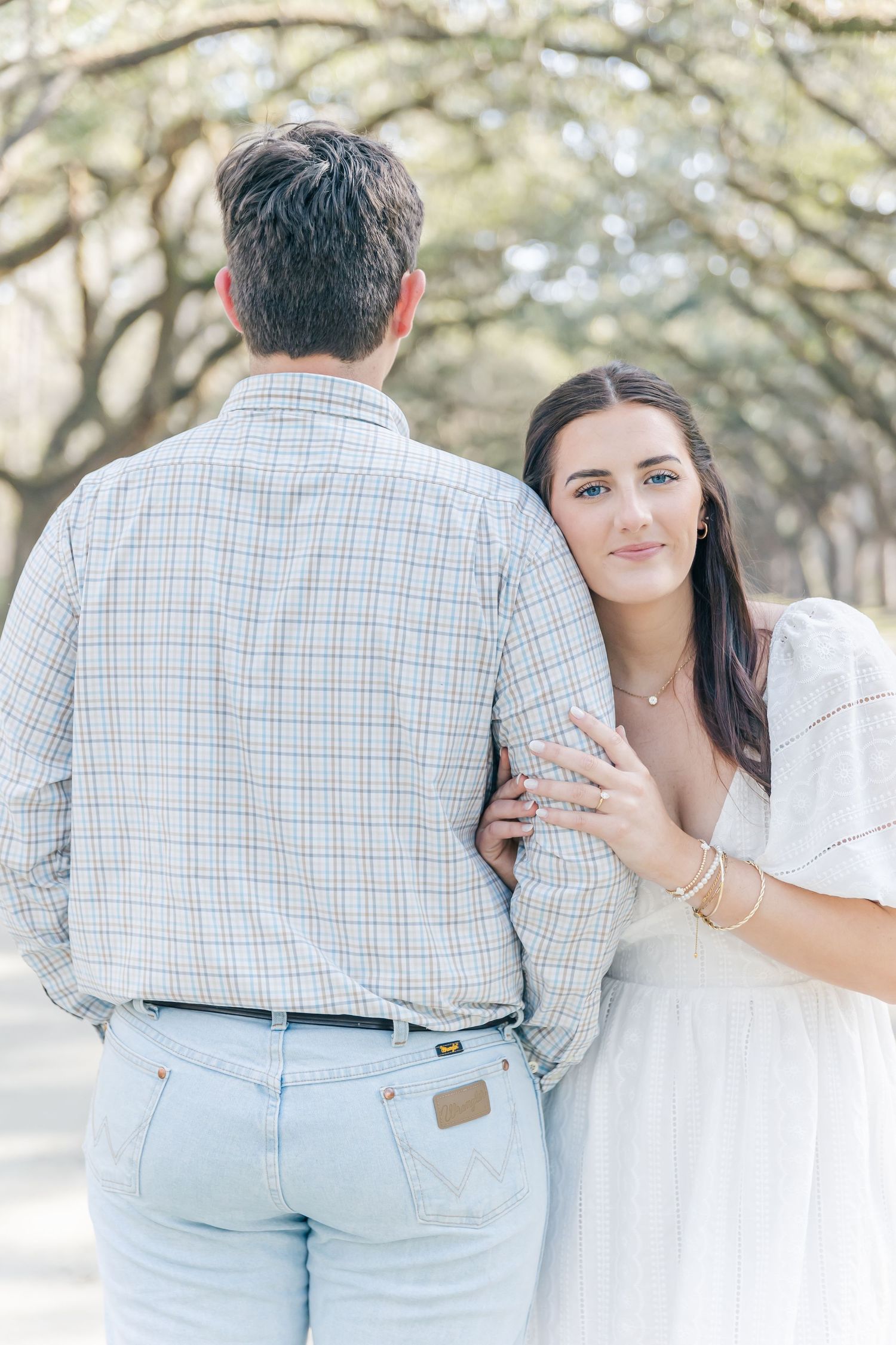 A couple poses together showing engagement ring while standing under oak trees.
