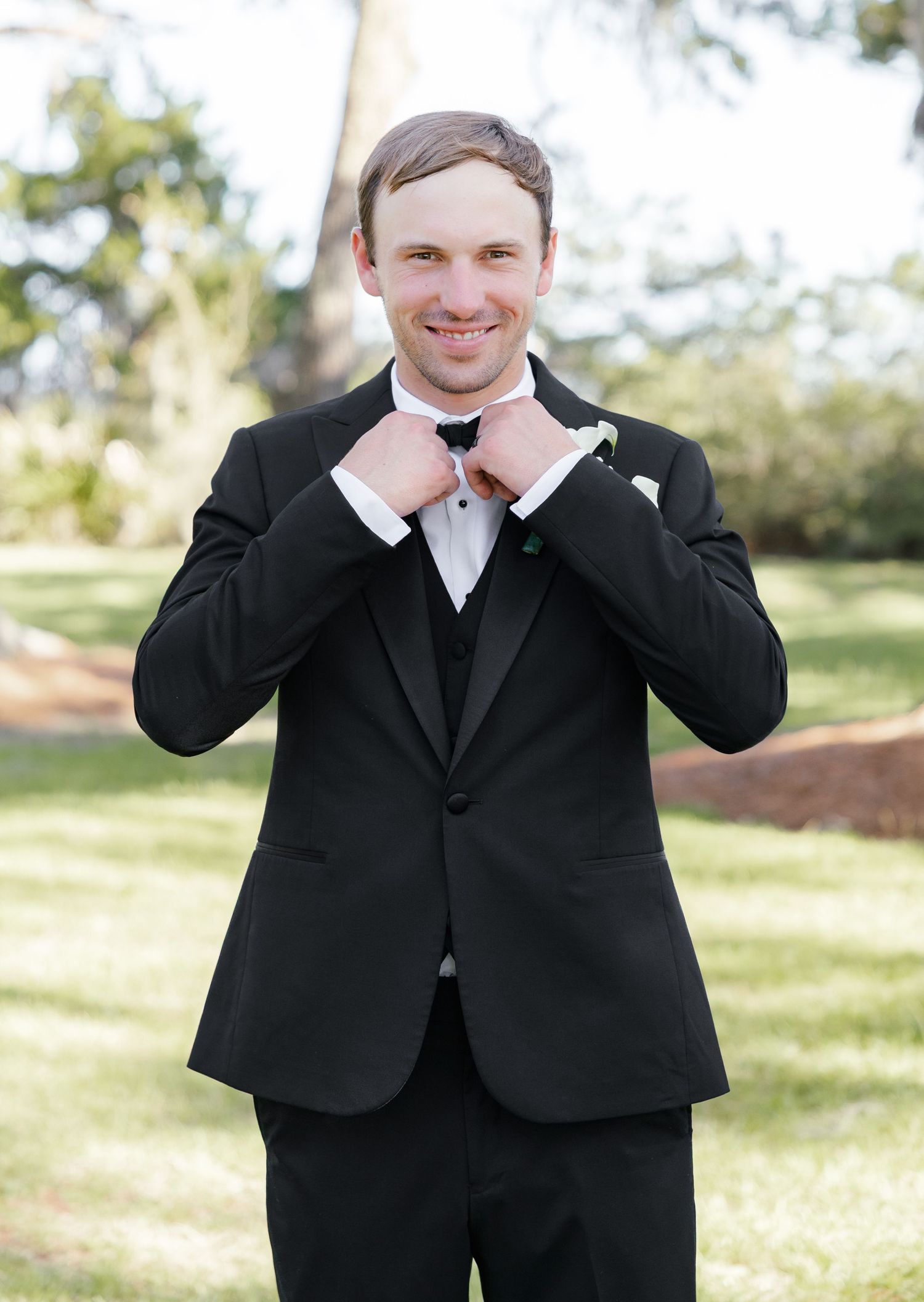 Groom in black tuxedo adjusting bow tie outdoors, smiling with white boutonniere on lapel.