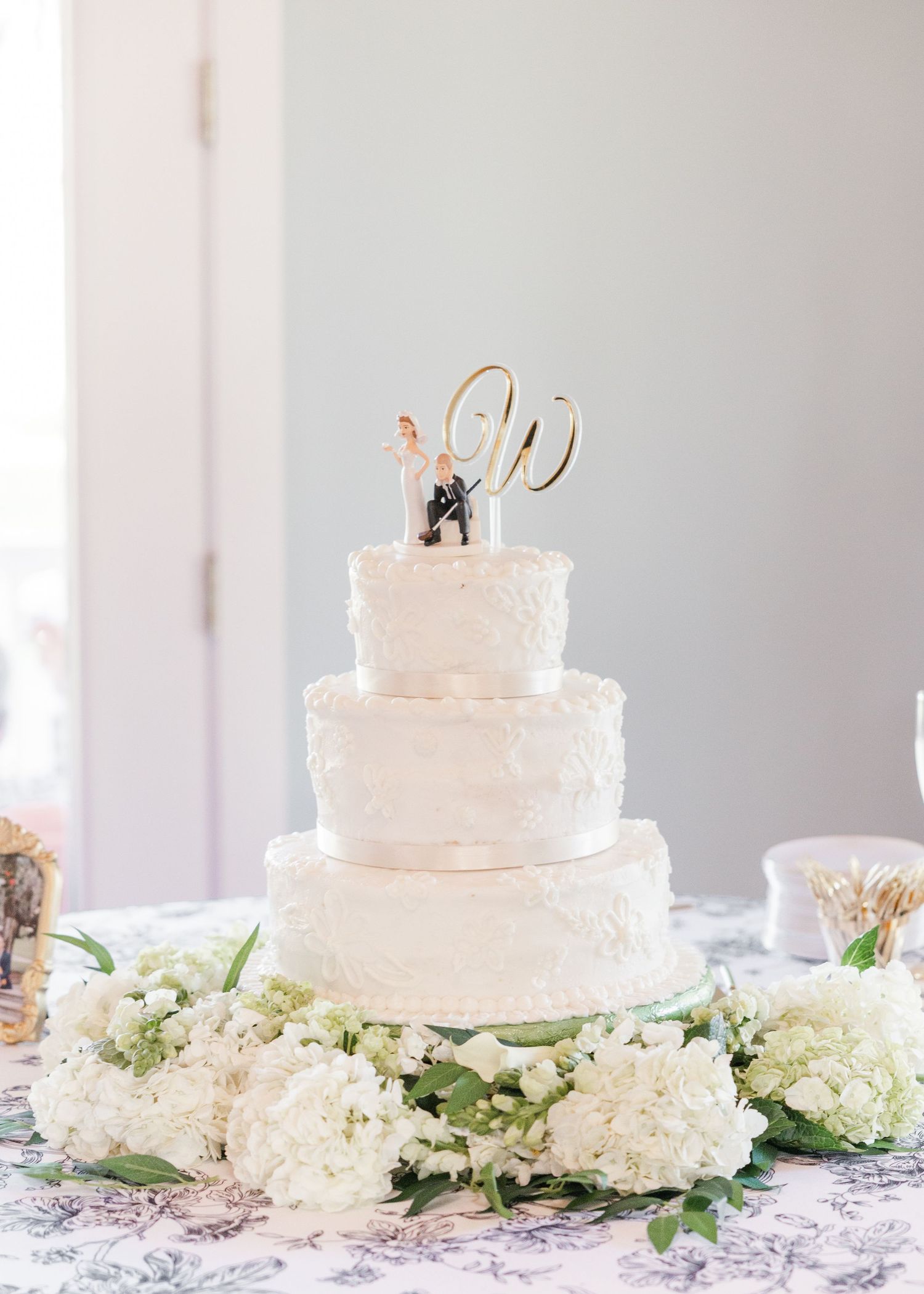 Three-tier white textured wedding cake with bride and groom figurine topper, gold W monogram, and white hydrangea floral base.
