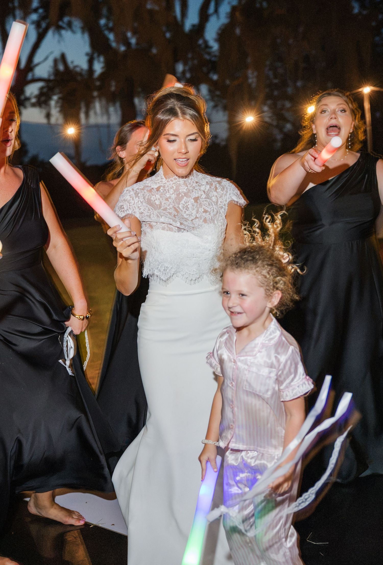 Bride in lace wedding gown celebrating with bridesmaids and flower girl holding glow sticks at night.