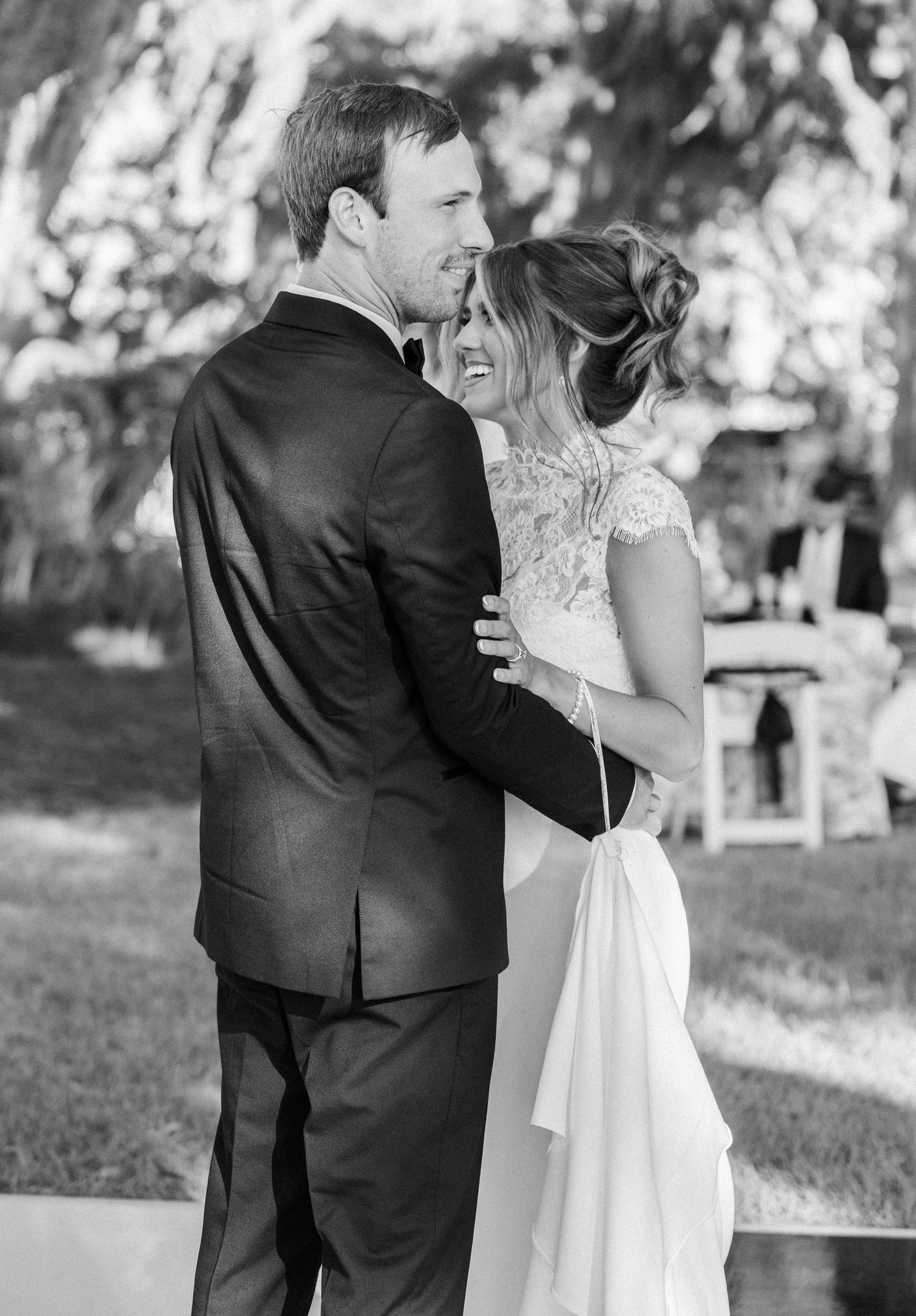 Black and white photo of a smiling bride in lace gown embracing a groom in a dark suit at an outdoor wedding.