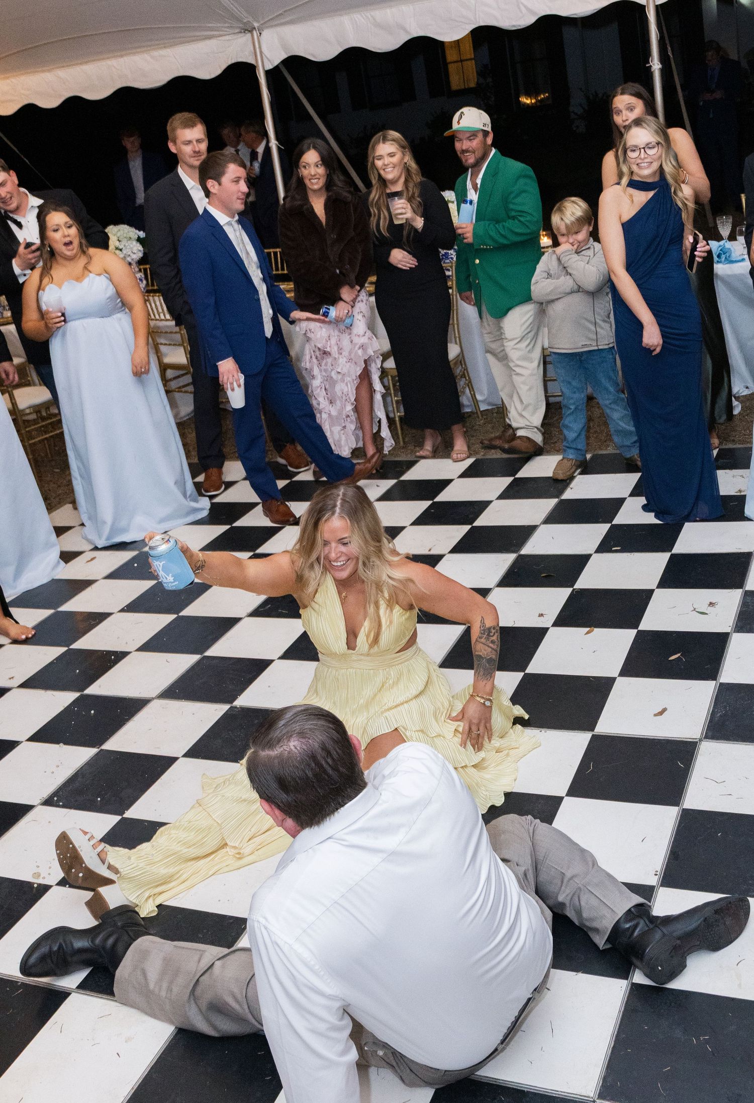 Wedding guests laugh as two people playfully dance on the black and white checkered reception dance floor.
