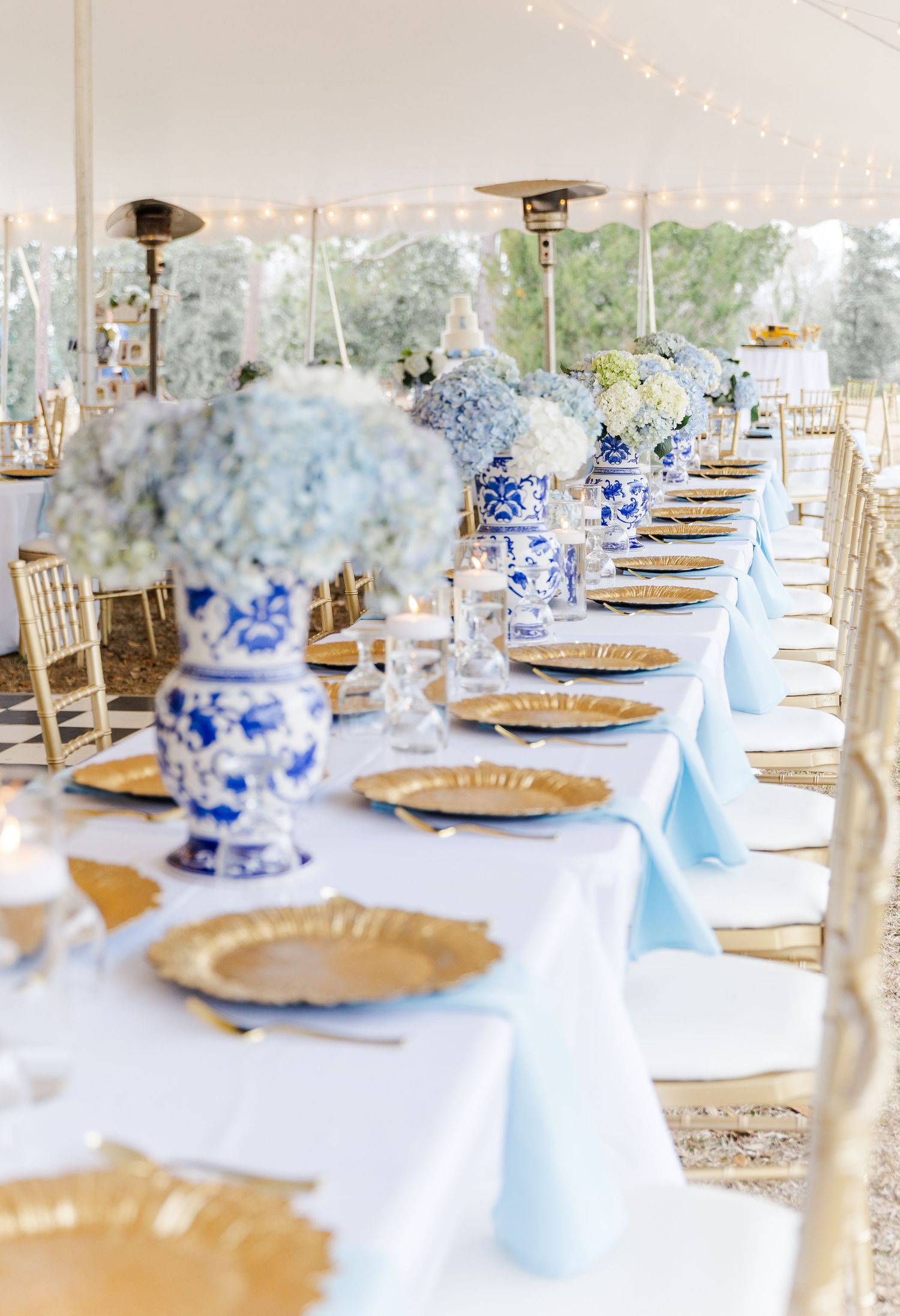 Long wedding reception table with blue hydrangeas in chinoiserie vases, gold chargers, and candles.