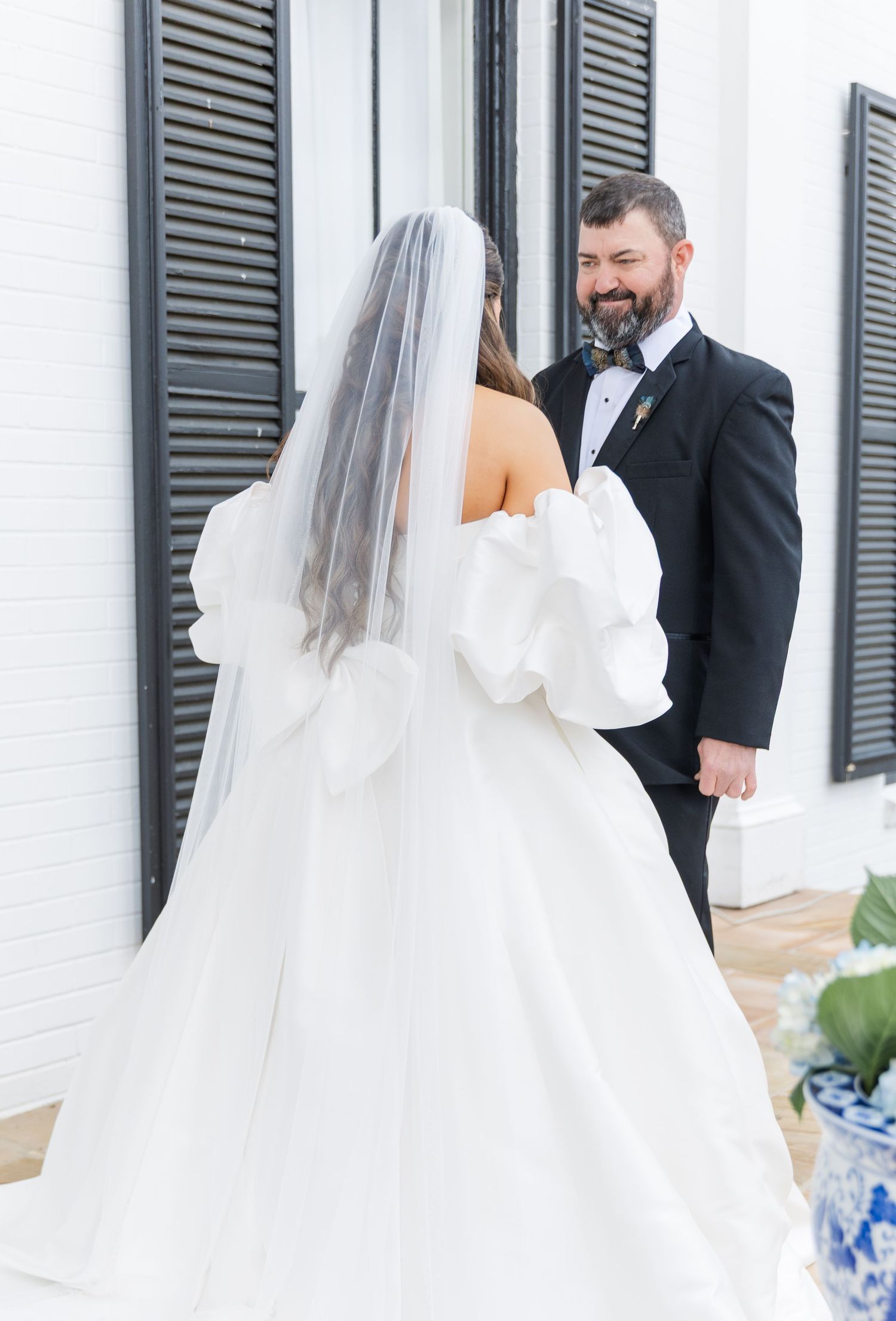 Bride in white ballgown with puff sleeves and veil shares first look with groom in black tuxedo outdoors.