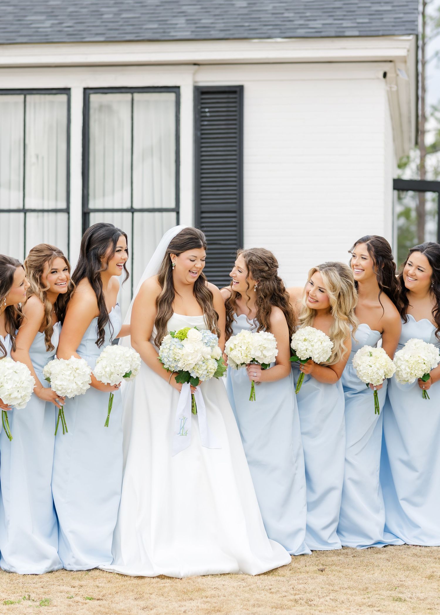 Bride and bridesmaids in light blue dresses laughing together holding white hydrangea bouquets.
