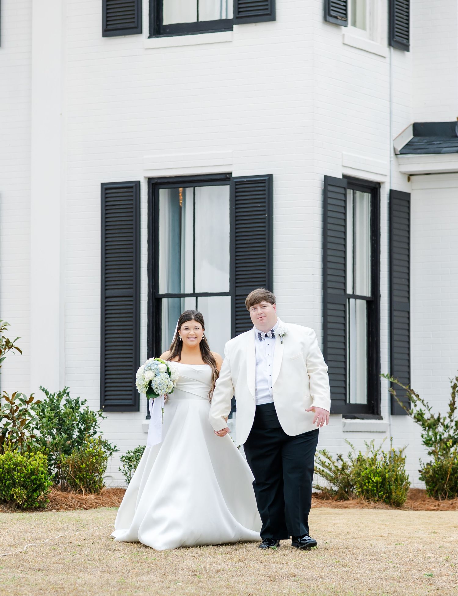 Bride and groom standing in front of white plantation-style venue with black shuttered windows.