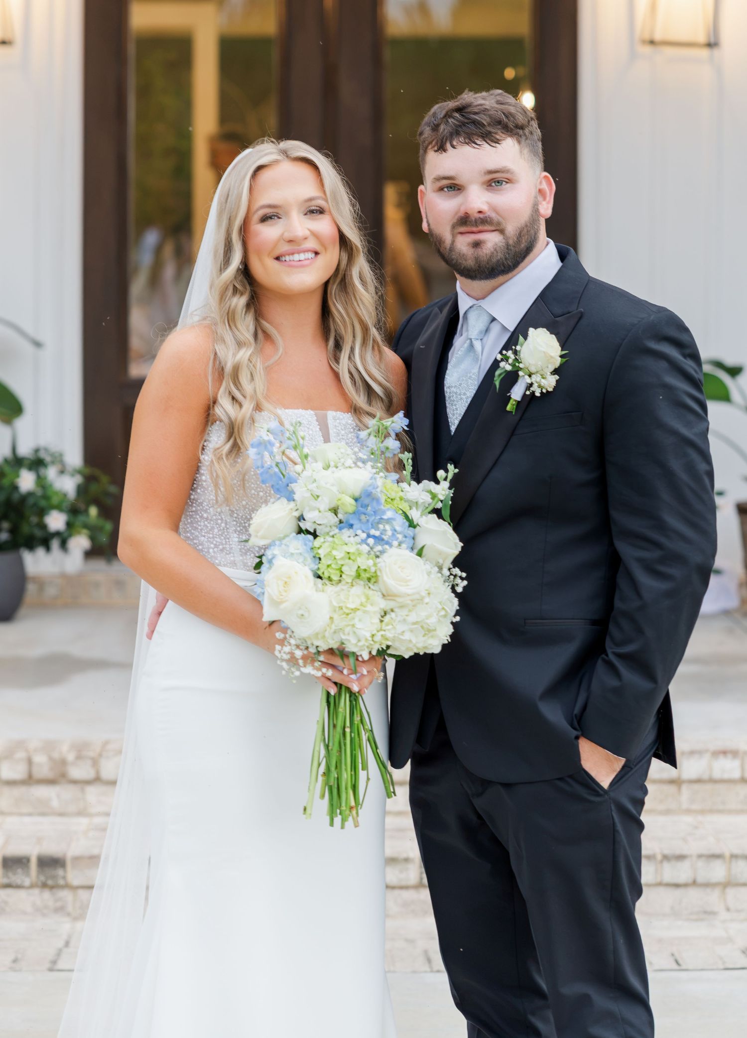 Smiling bride and groom posing together outdoors, bride holding blue and white hydrangea bouquet.