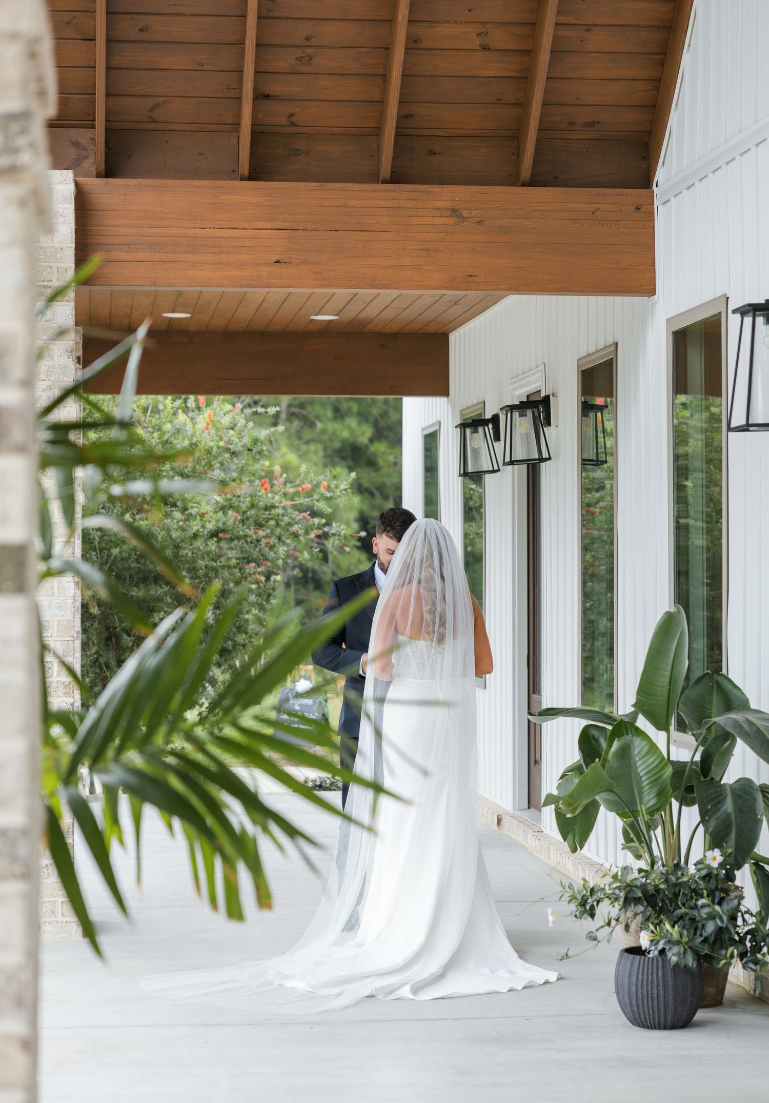 Romantic moment of bride and groom sharing first look on white farmhouse venue porch surrounded by tropical plants.