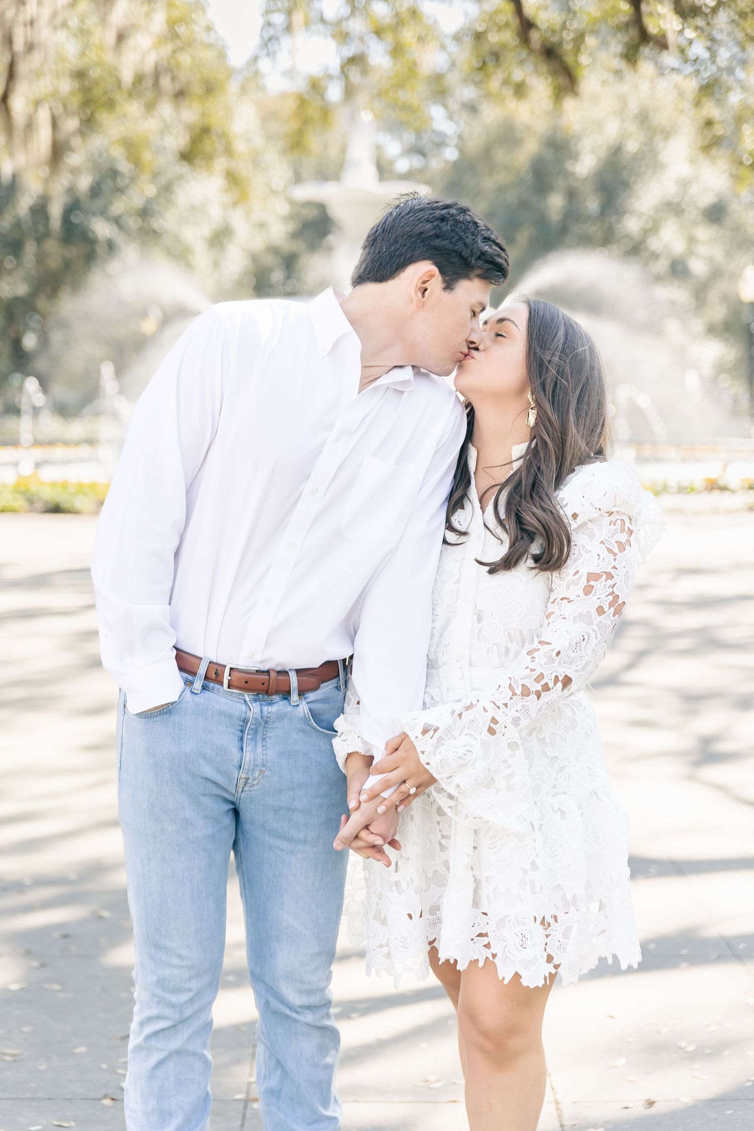 A romantic couple outdoors embraces in soft sunlight, with her wearing a white lace dress and him in jeans and white shirt.