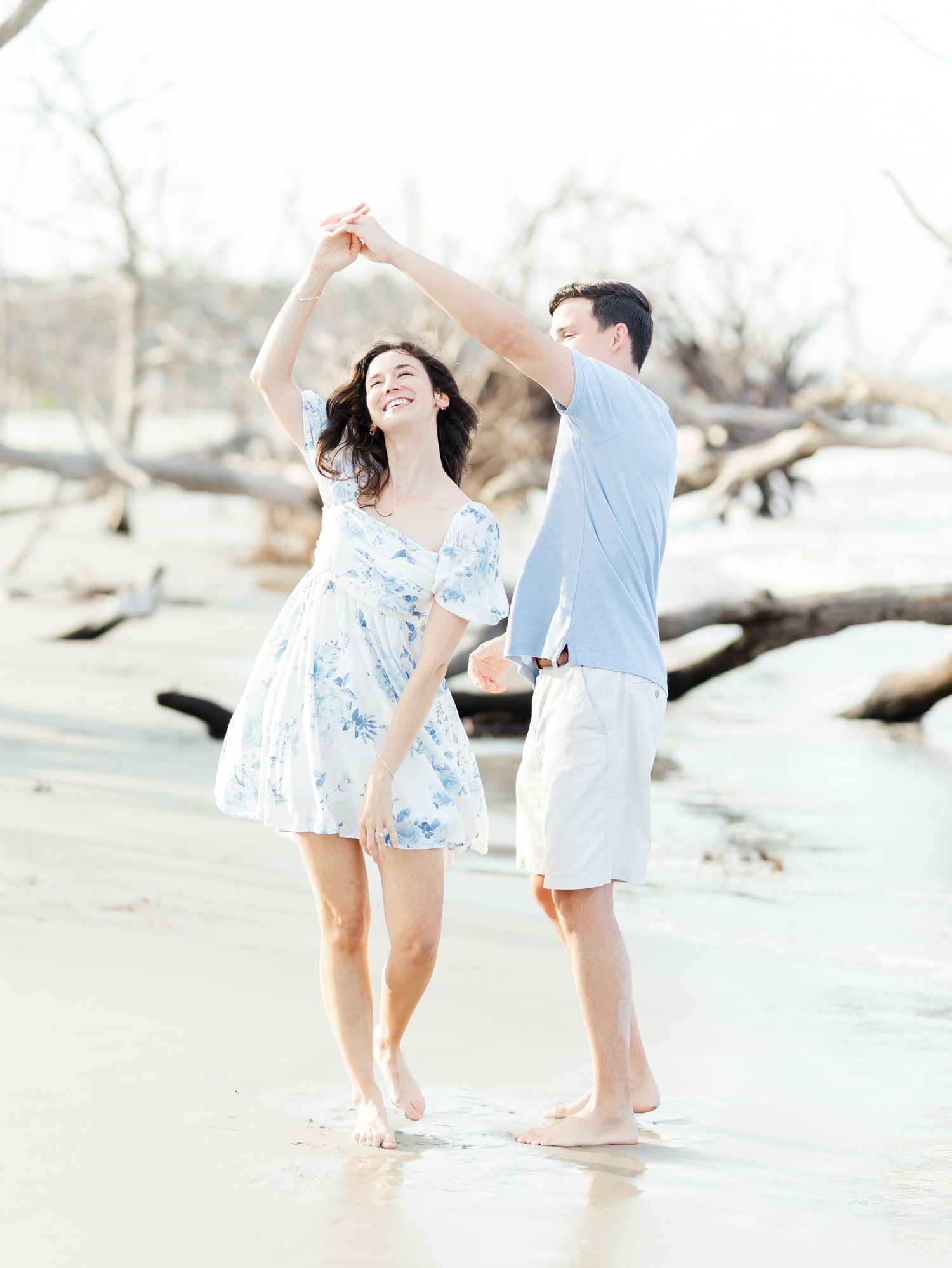 A couple in white and blue outfits dance playfully together on a bright, sandy beach.