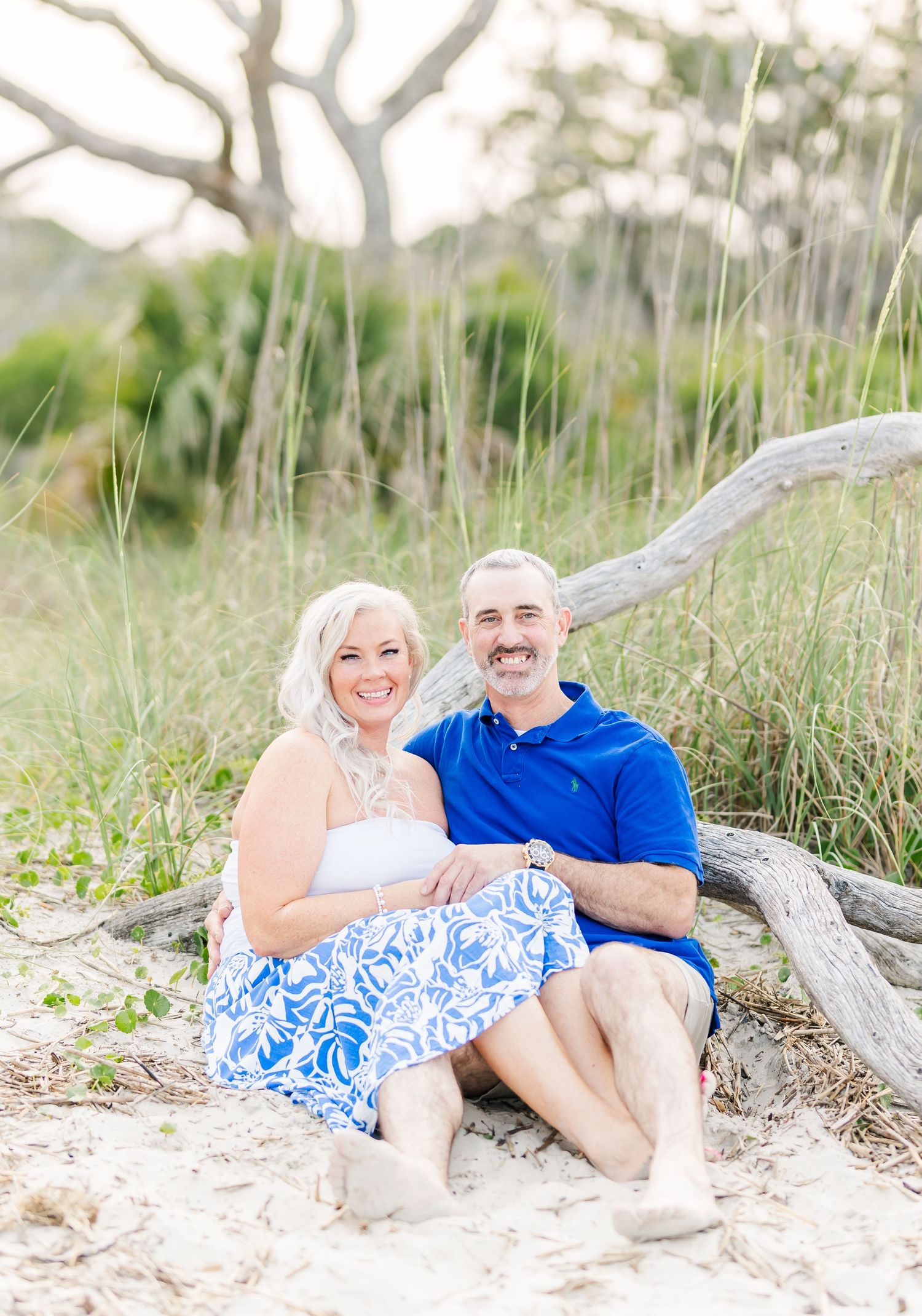 A couple sits together on driftwood at the beach surrounded by sea grass on a bright sunny day.
