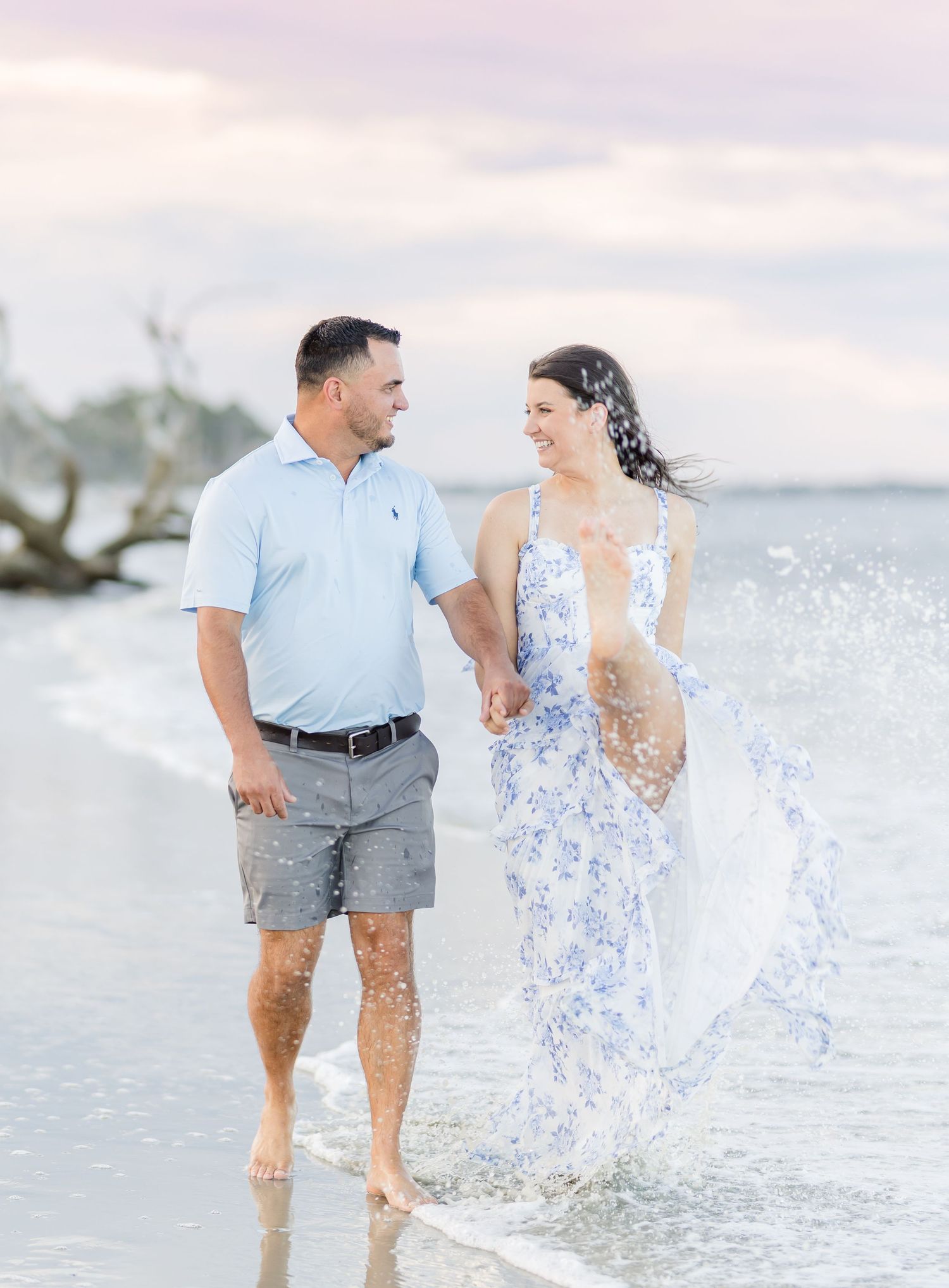 A couple in coordinating blue and white outfits splashes playfully along the shoreline at sunset.