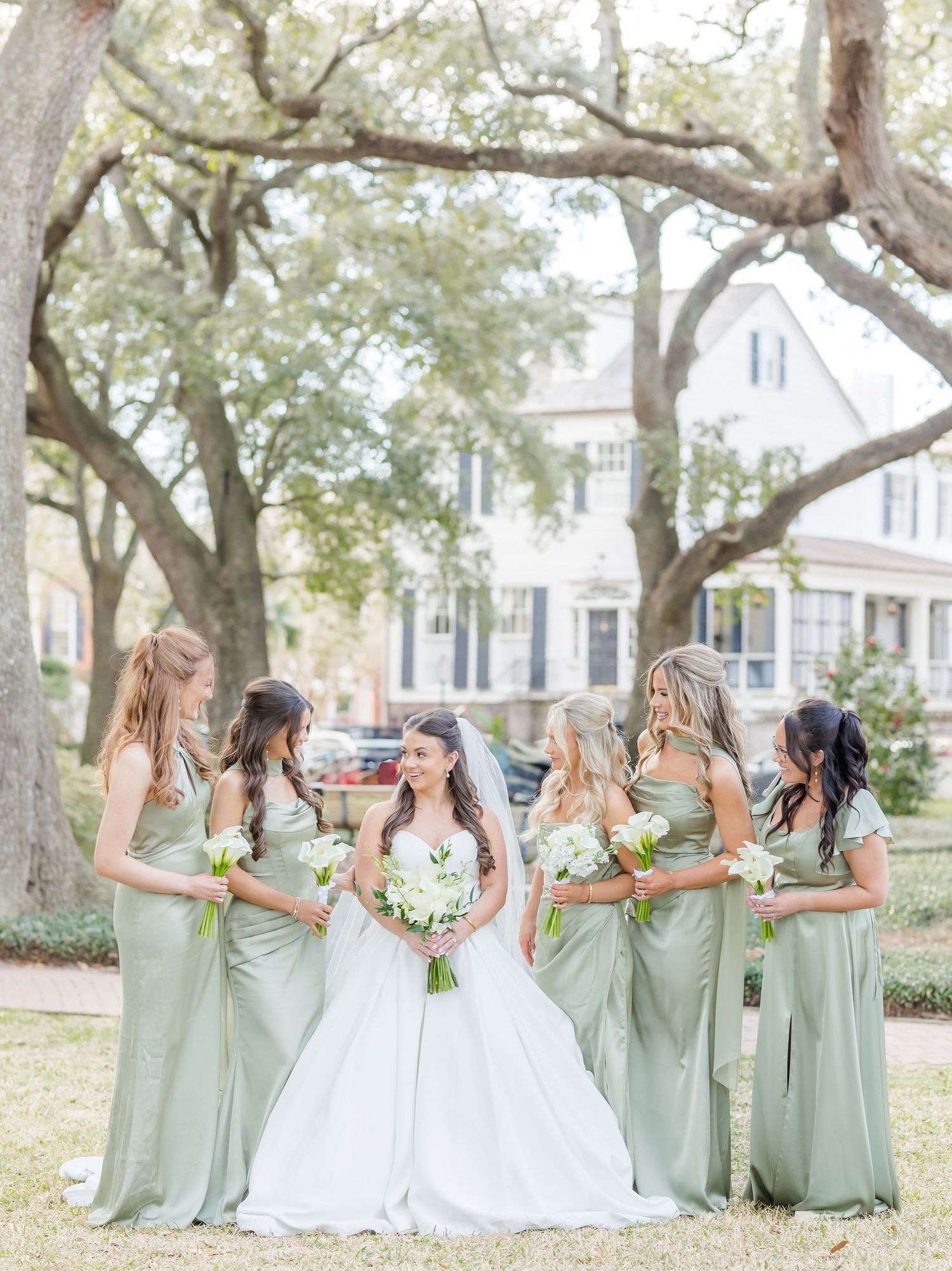 Bride in white gown stands with five bridesmaids in sage green dresses holding white bouquets outdoors.