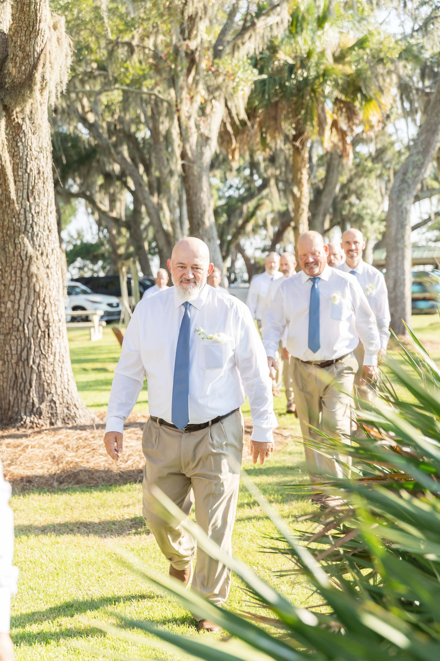 Groomsmen in white shirts and blue ties walk through a scenic park with Spanish moss draped trees.