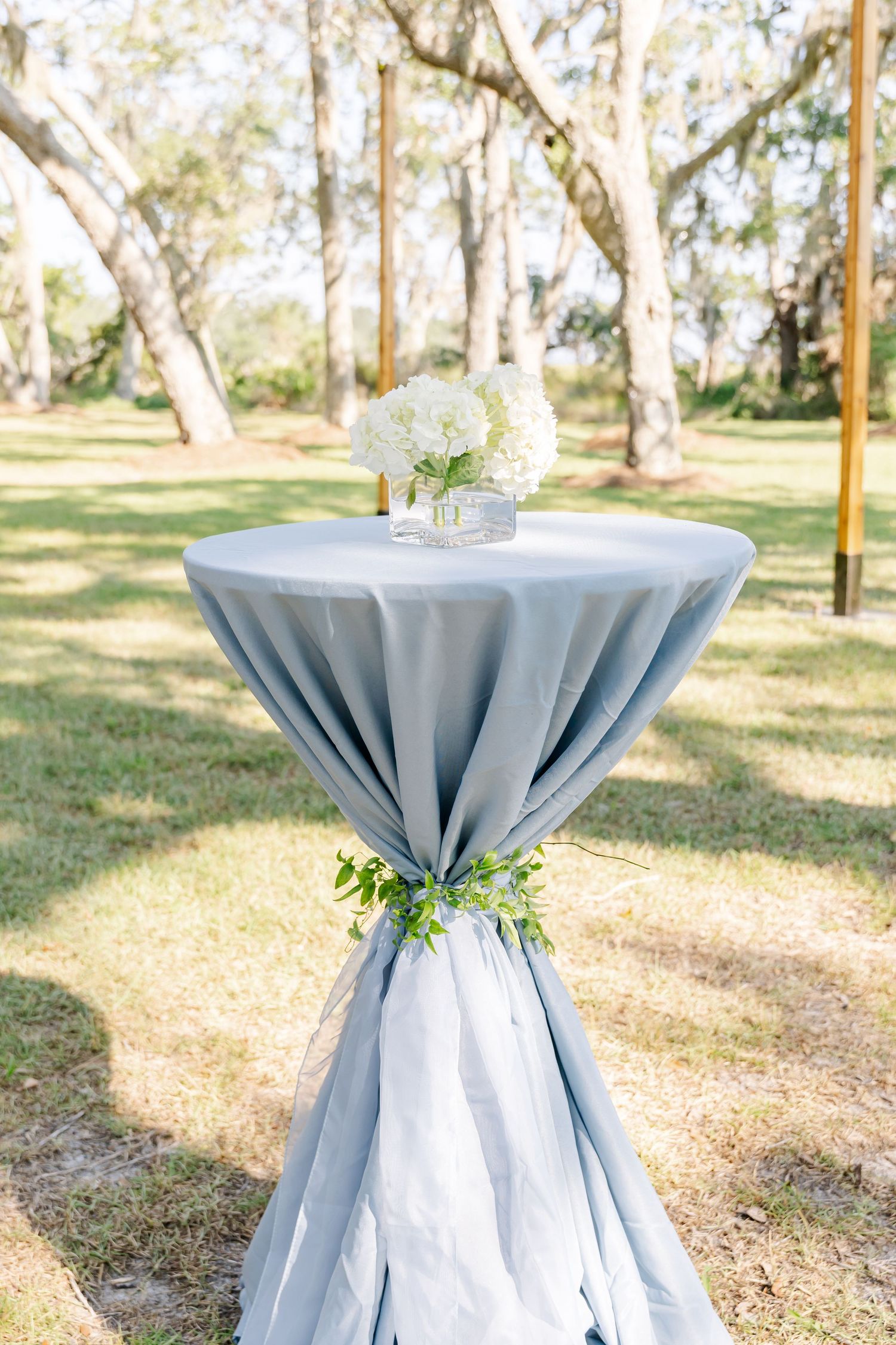 Light blue cocktail tables with white linens and floral arrangements set up outdoors among trees.