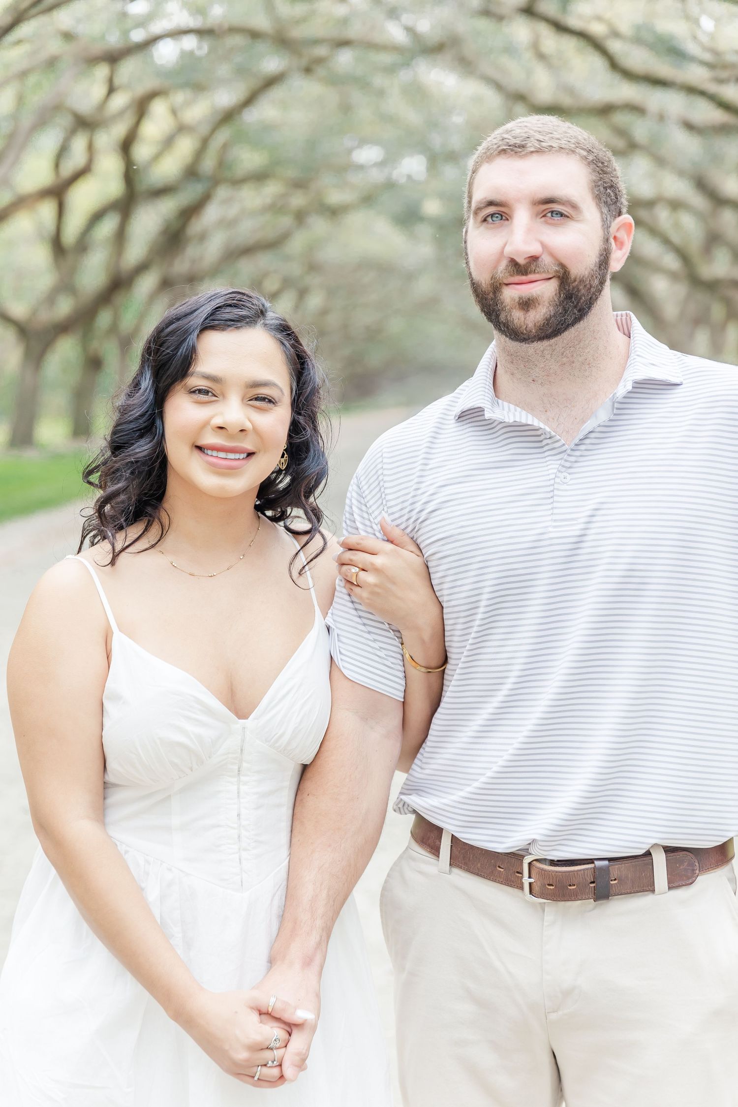 Happy couple in white and striped attire pose beneath canopy of oak trees.