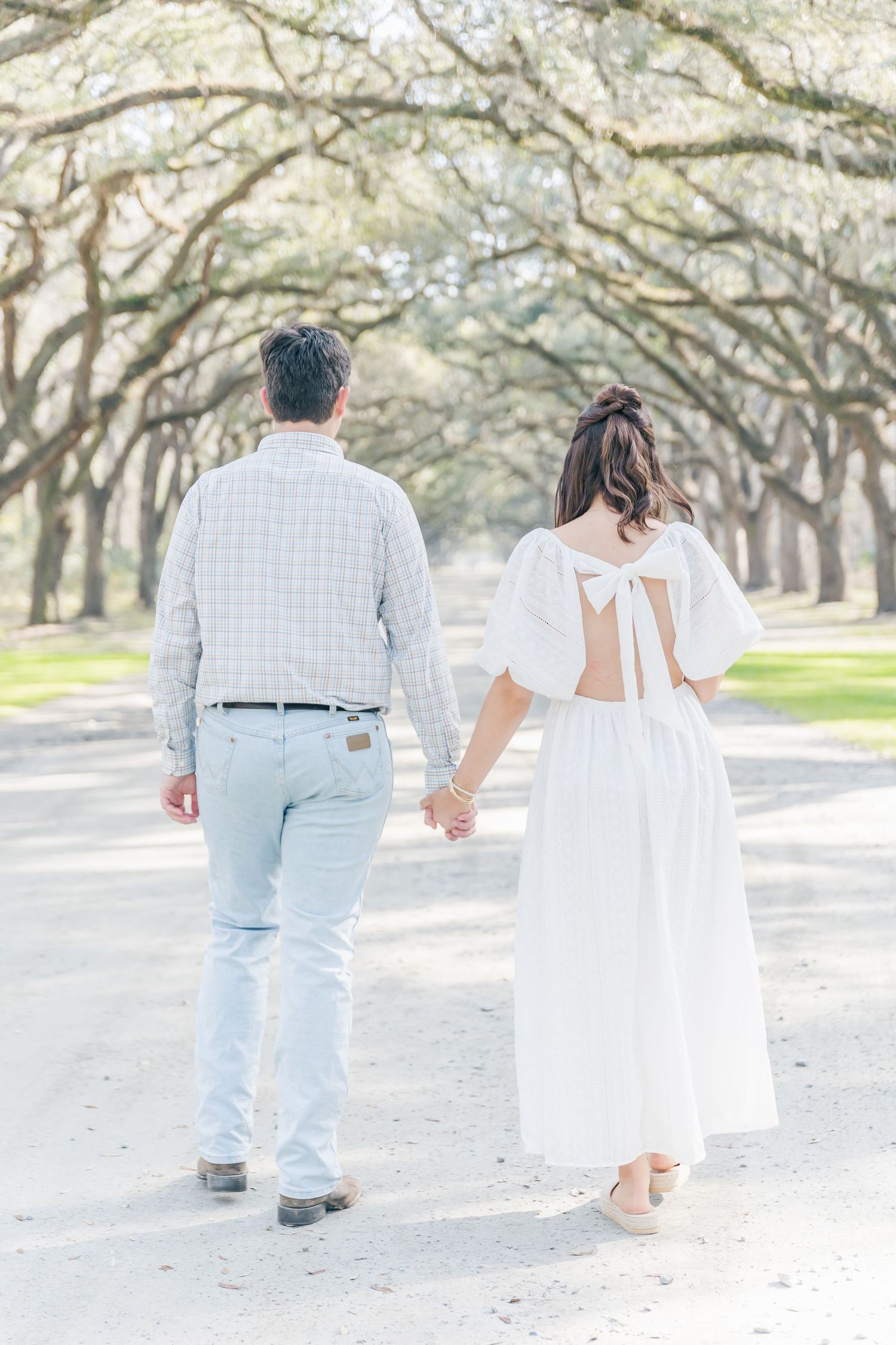 Back view of a couple strolling down an oak-lined avenue, with the woman's dress featuring a bow detail.