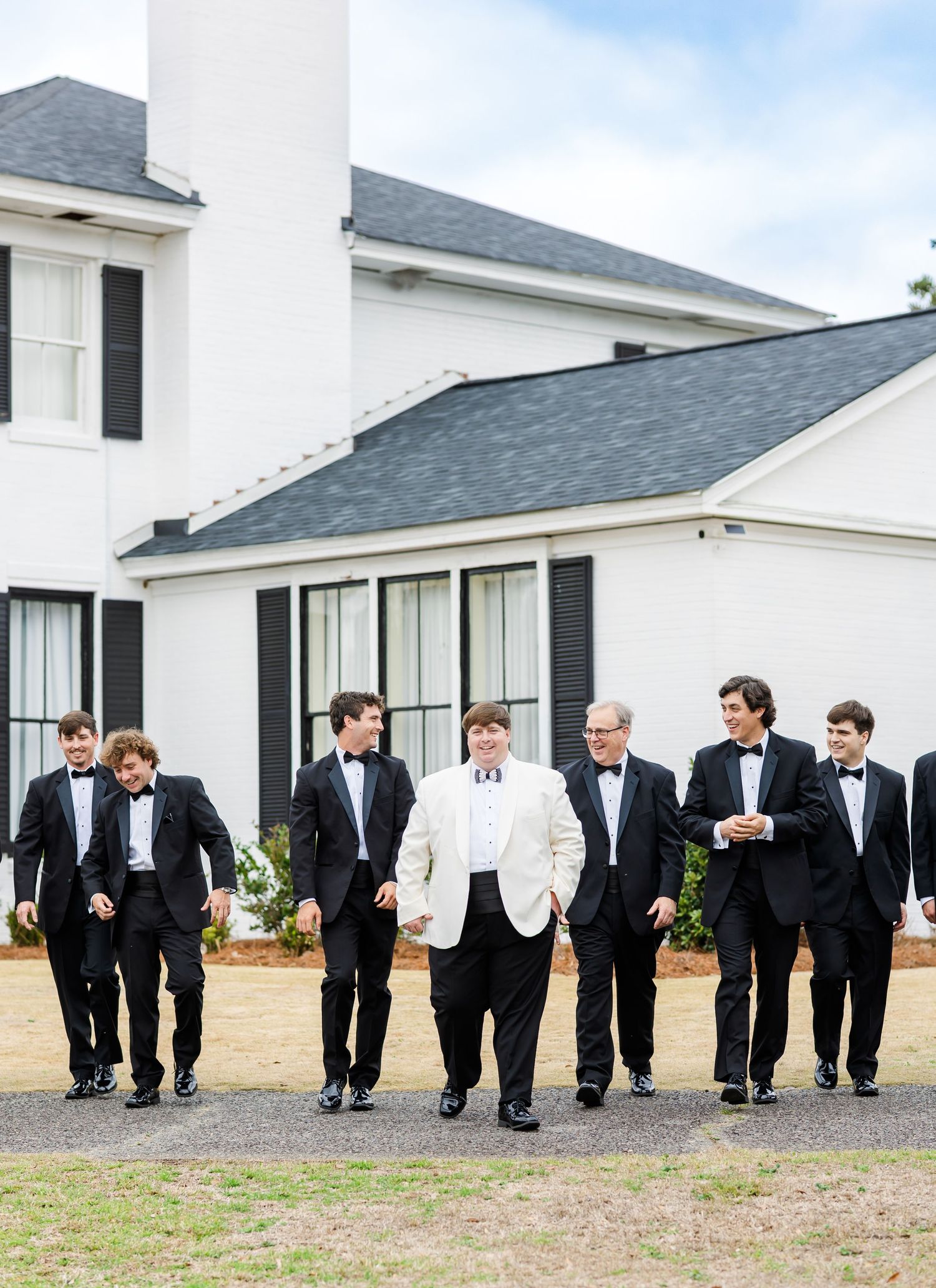 Groom in white jacket walks with groomsmen in black tuxedos outside a white Southern estate venue.