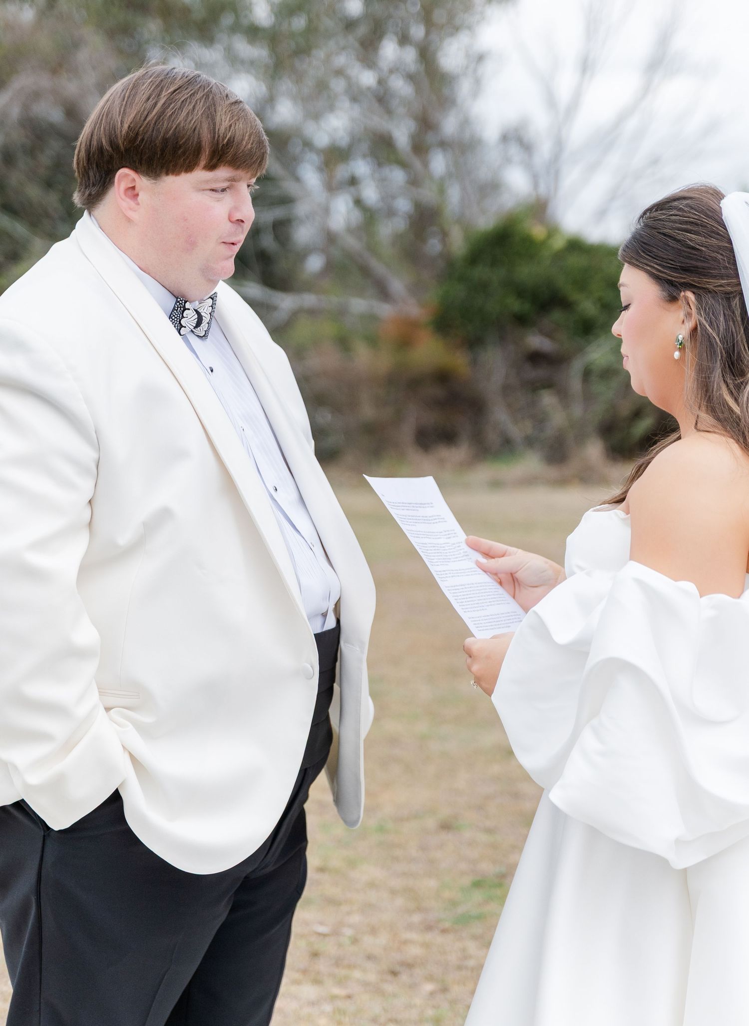 Bride in white off-shoulder gown reads vows to groom in ivory tuxedo jacket outdoors at wedding ceremony.