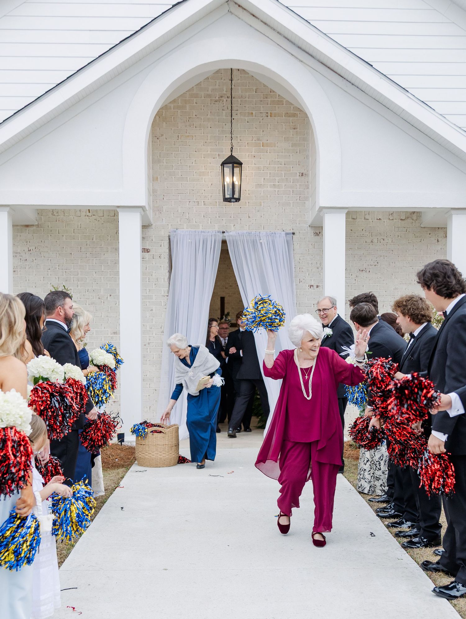 Elderly woman in magenta suit exits white brick chapel as wedding guests wave colorful pom-poms in celebration.