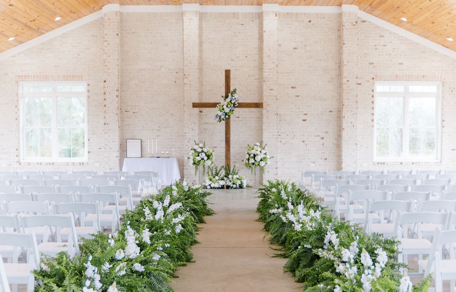 Serene white chapel wedding ceremony with wooden cross, white floral aisle garlands, and rows of white chairs.