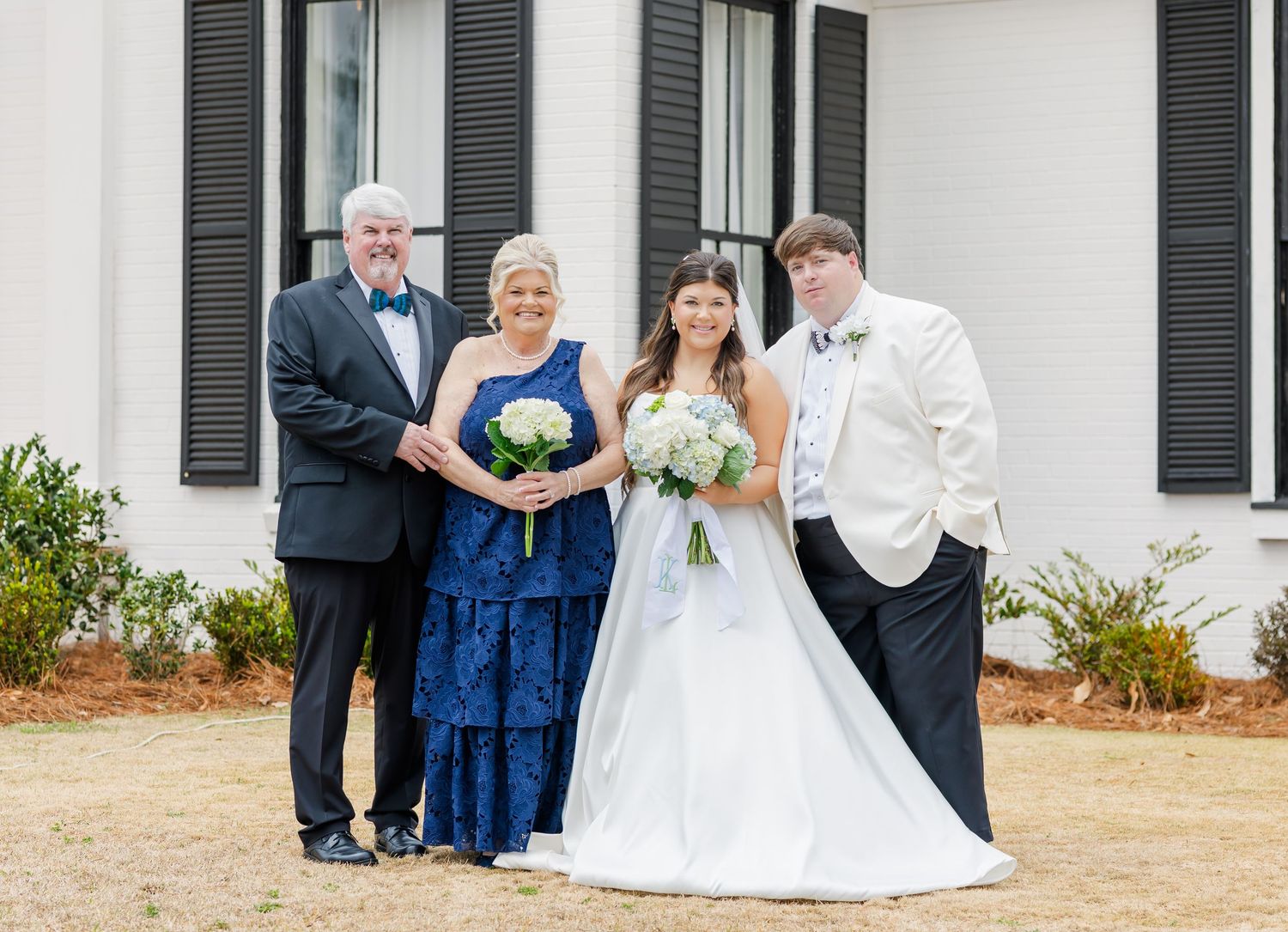 Bride in white gown poses with family including parents and sibling outside a white wedding venue with black shutters.