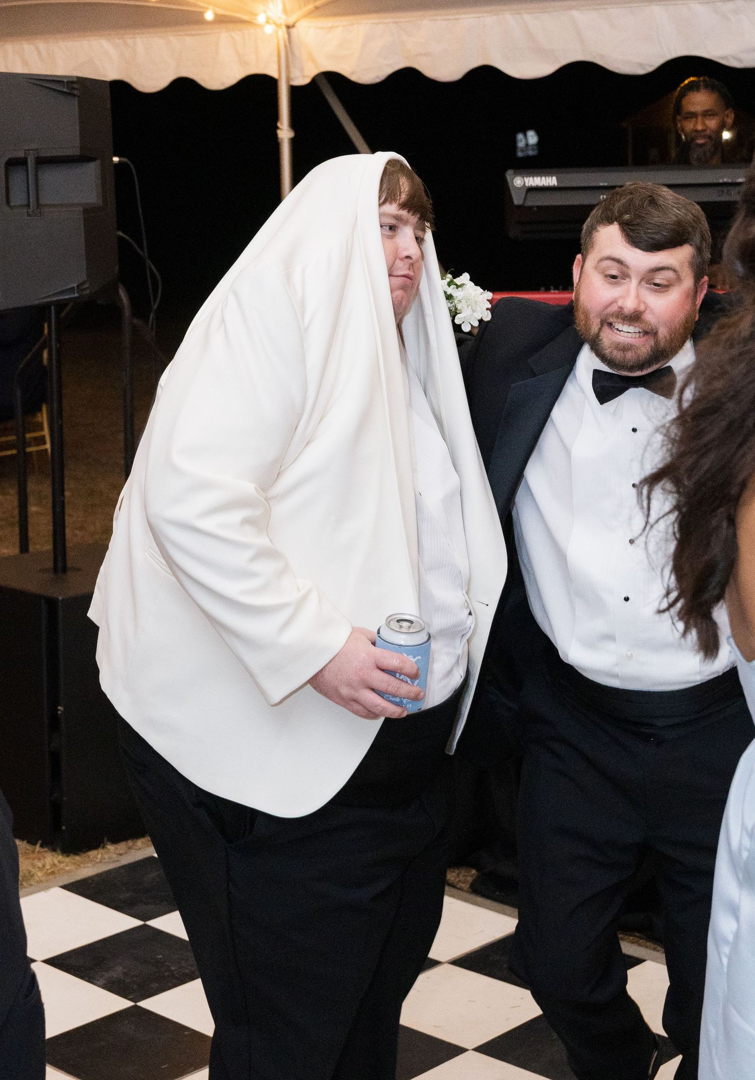 Man humorously draped in white tablecloth dances with tuxedo-clad guest on checkered dance floor at wedding.