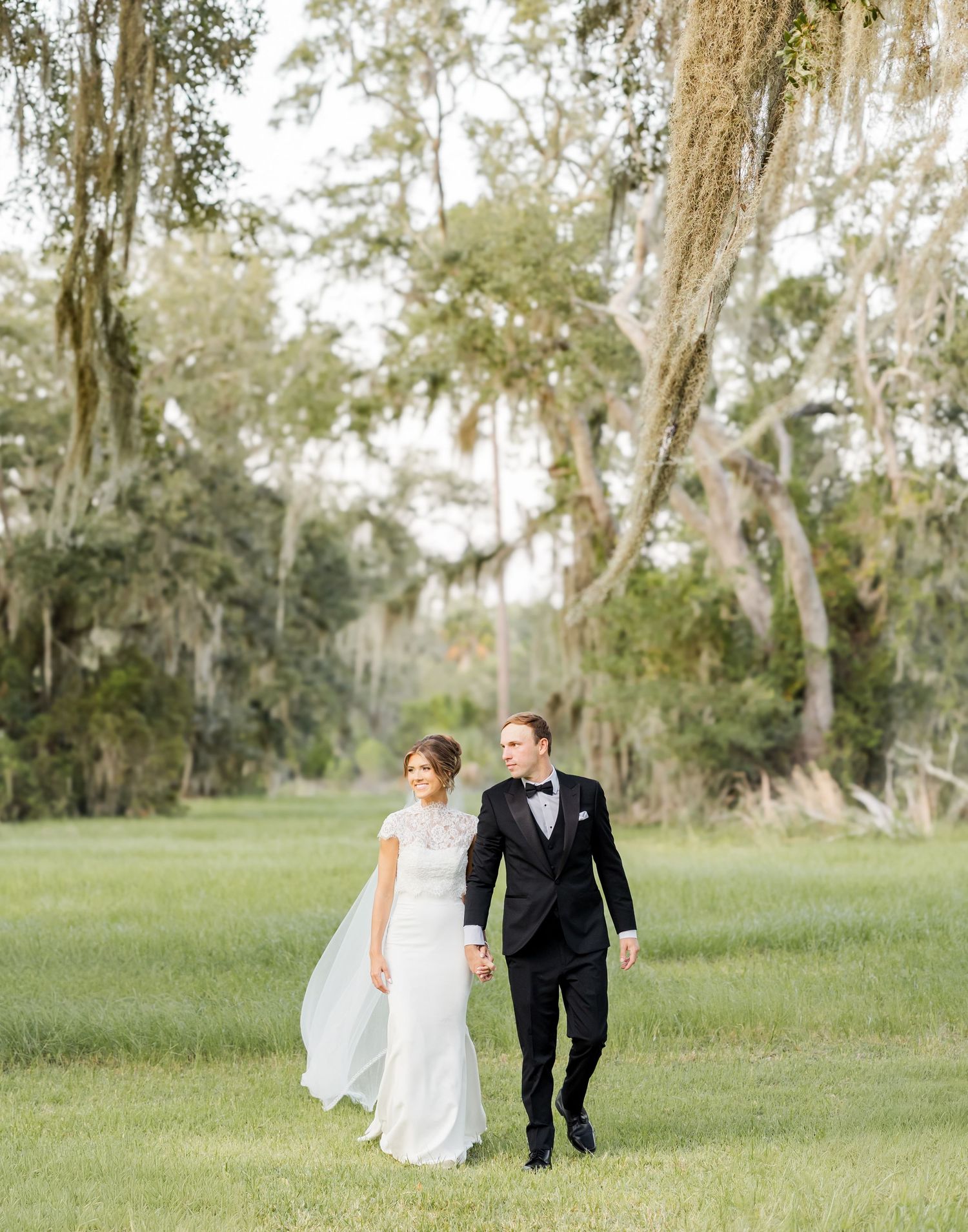 Bride and groom walking hand in hand through a lush green field with moss-draped oak trees at their wedding.