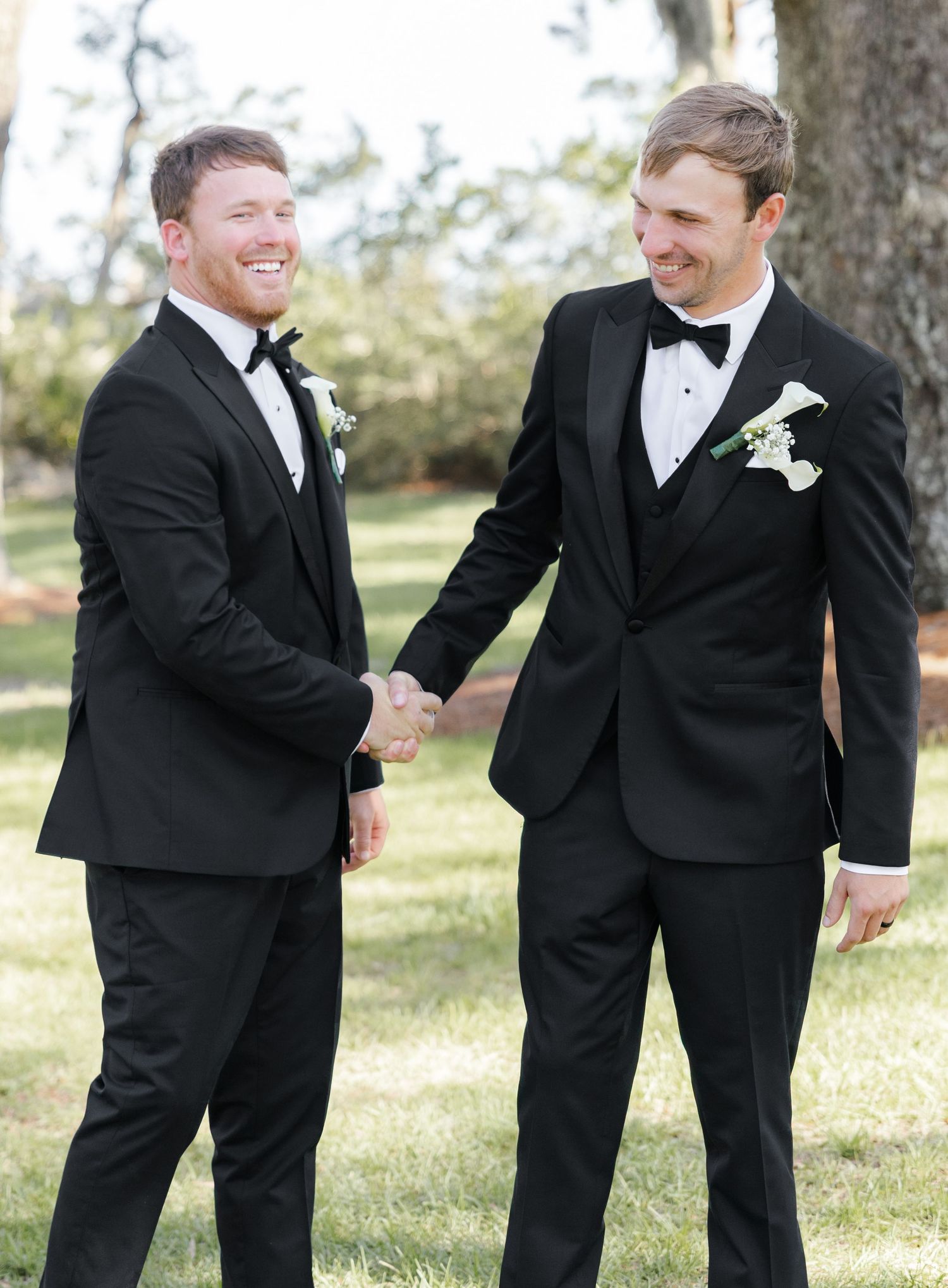 Two groomsmen in black tuxedos with white boutonnieres share a handshake and smile outdoors at a wedding.