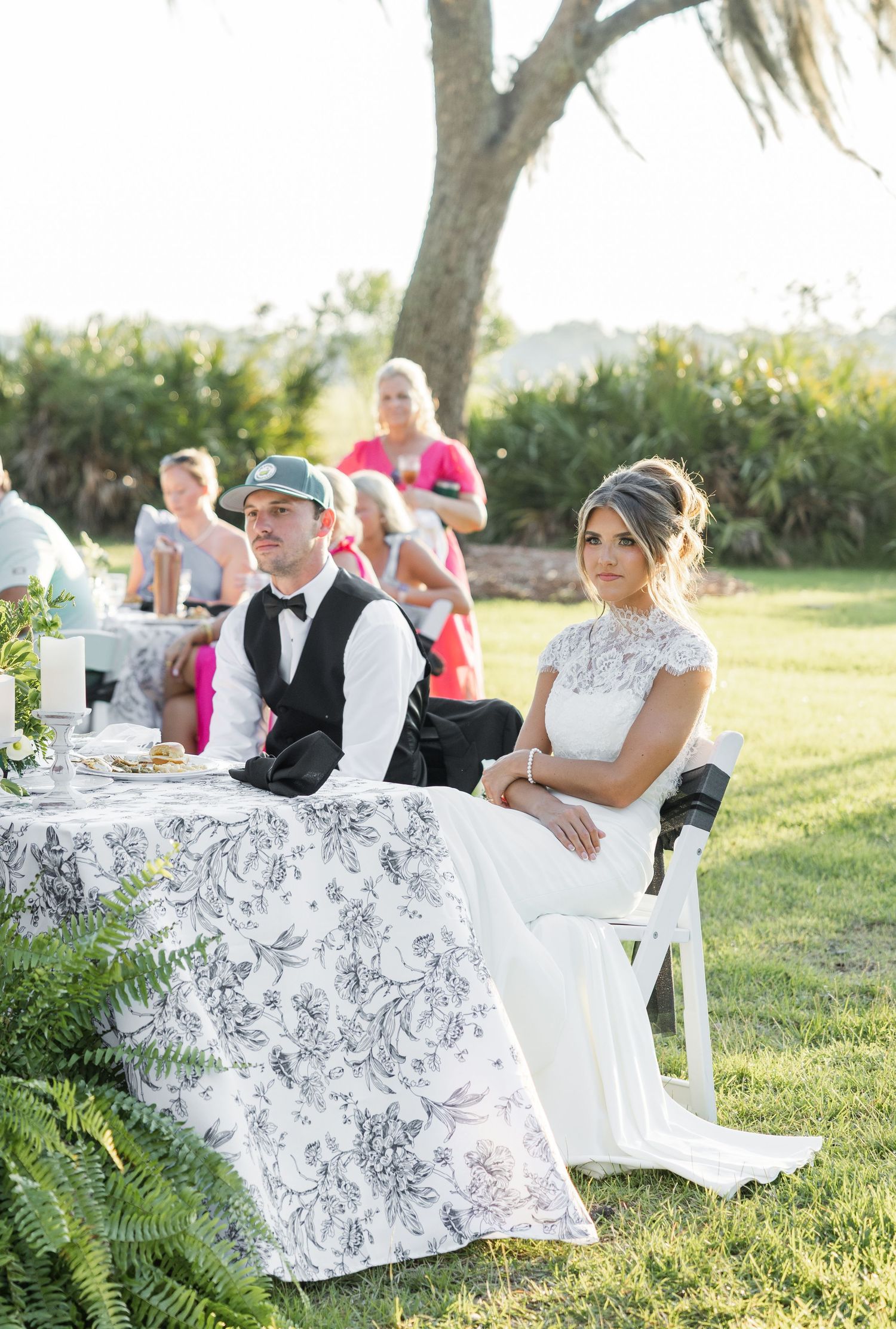 Bride in lace gown and groom in bow tie seated at floral tablecloth reception table outdoors at golden hour.