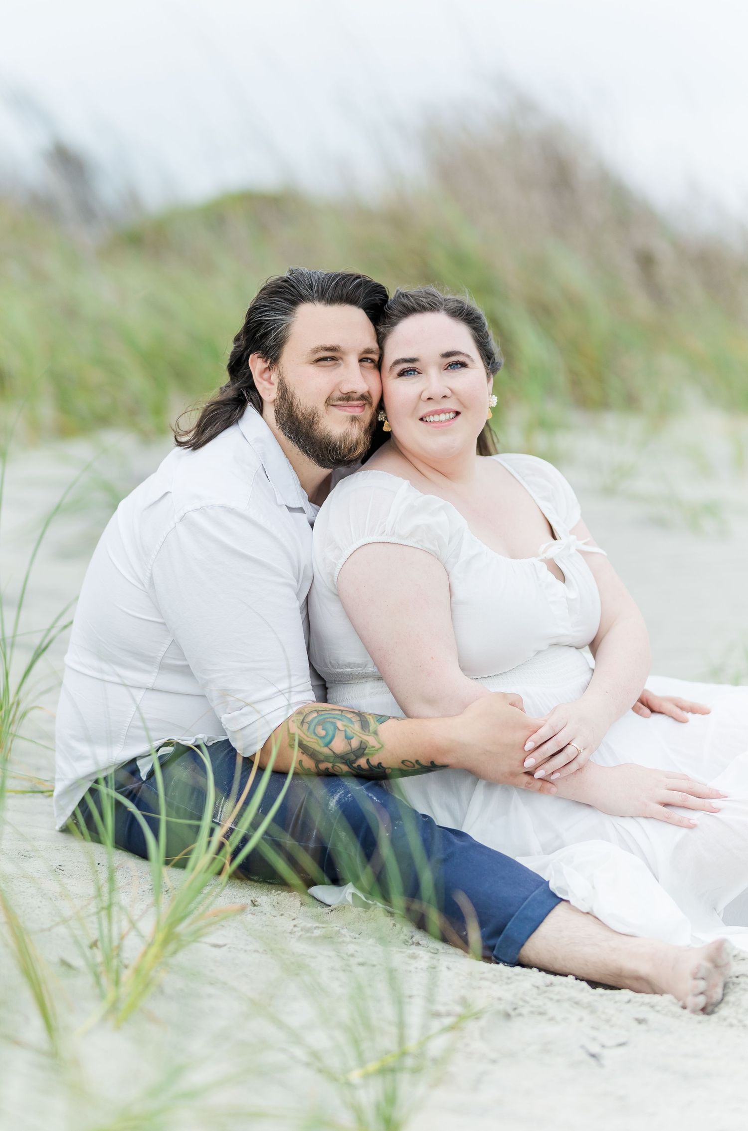 Couple sits together on beach dunes surrounded by sea grass in soft light.