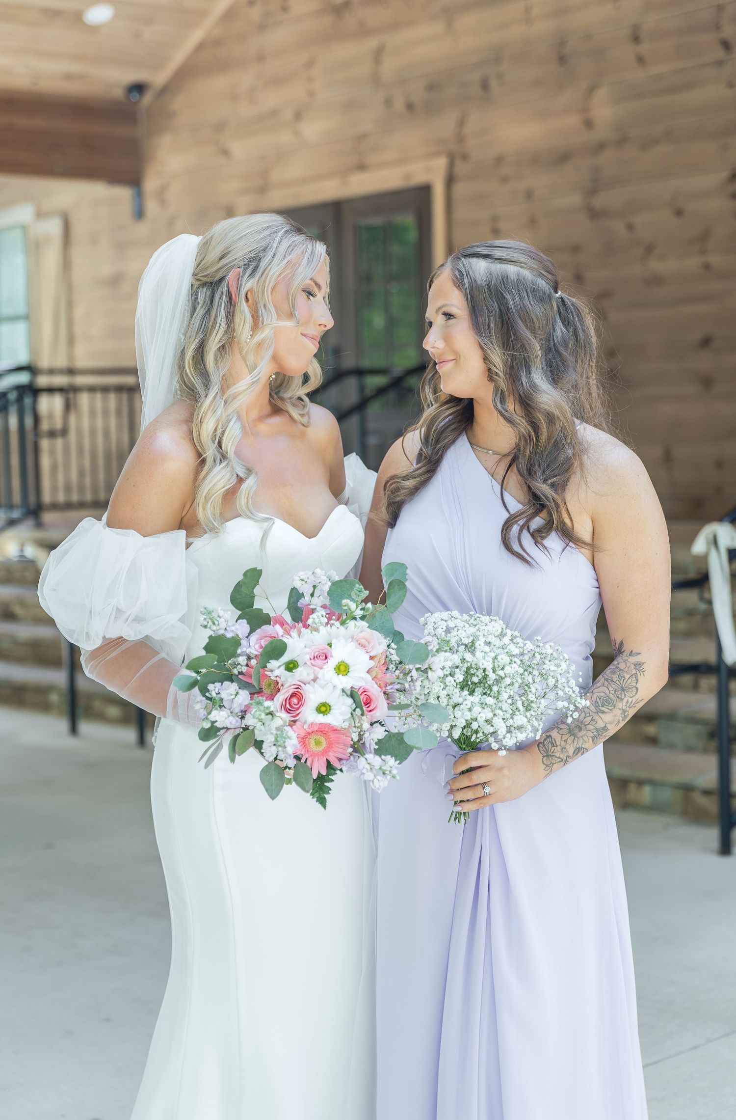Two women in bridal party attire smile at each other while holding pink and white bouquets.