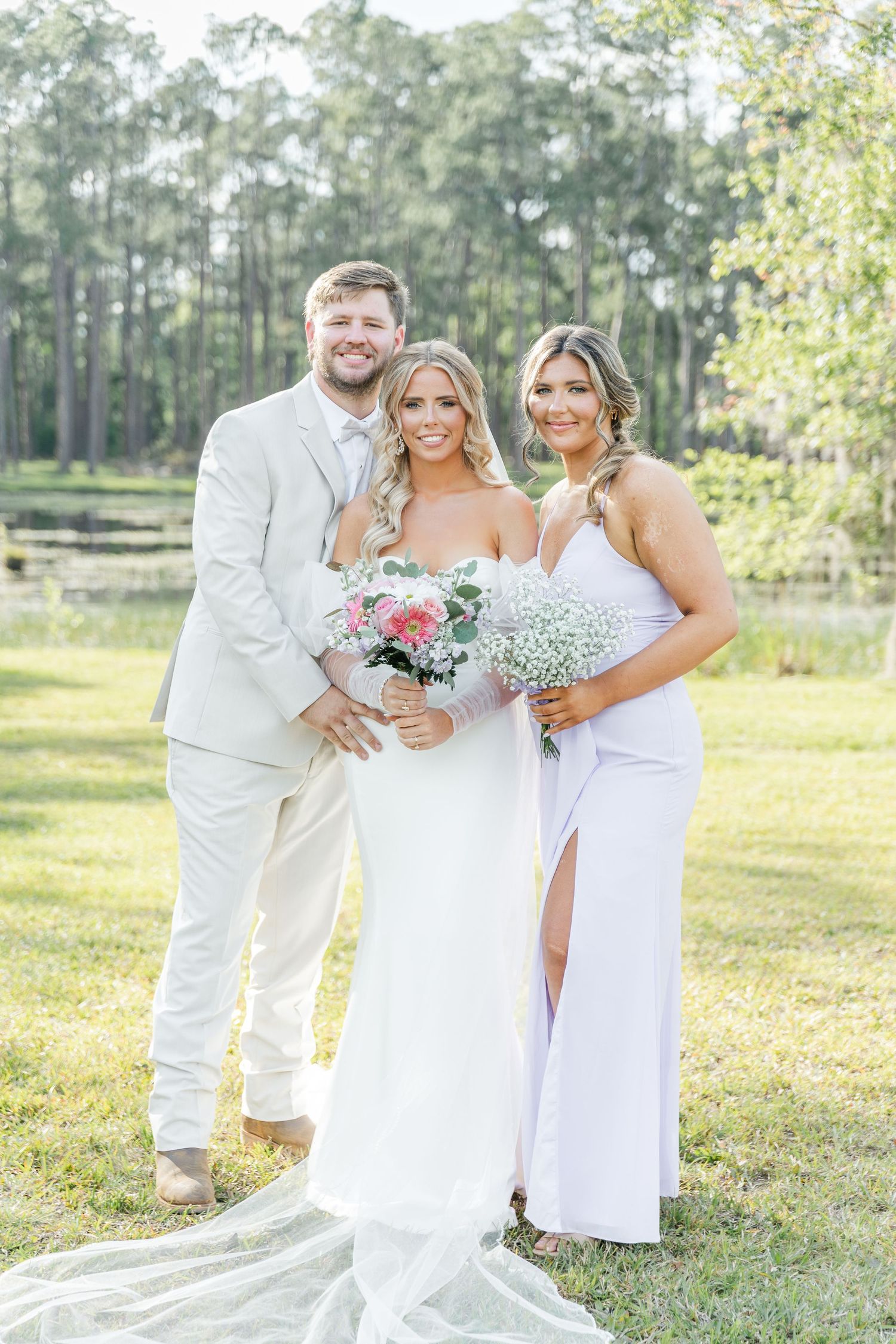 Wedding group photo in white attire with pink bouquet against wooded background.